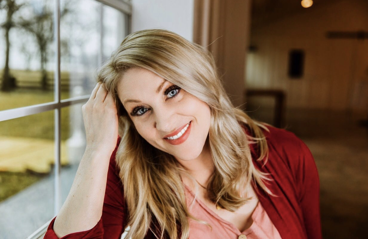 Woman with blonde hair smiling, wearing a red jacket, indoors near a window.