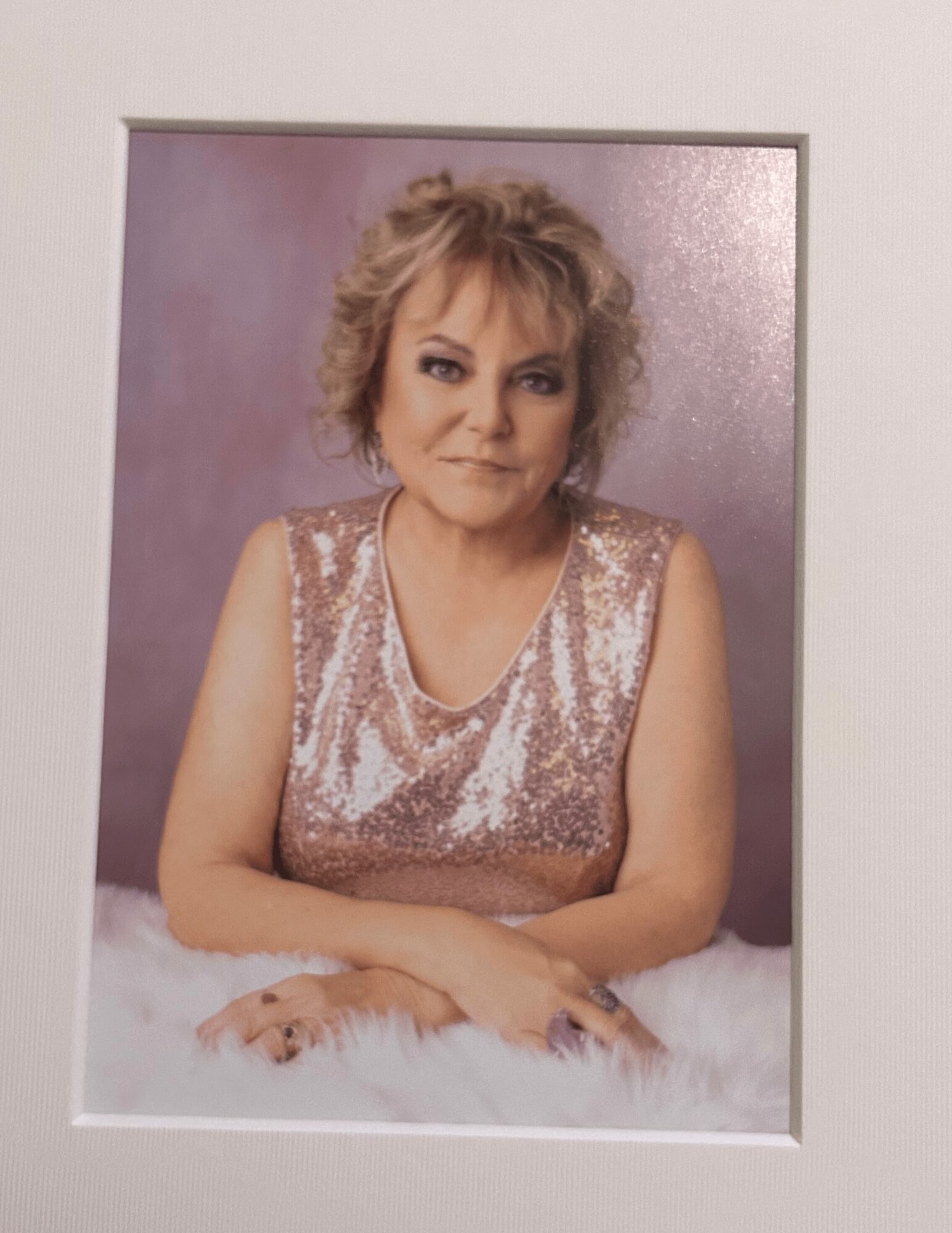 Woman with short curly hair wearing a sleeveless, sequined top, sitting with hands folded on a fluffy surface, smiling.