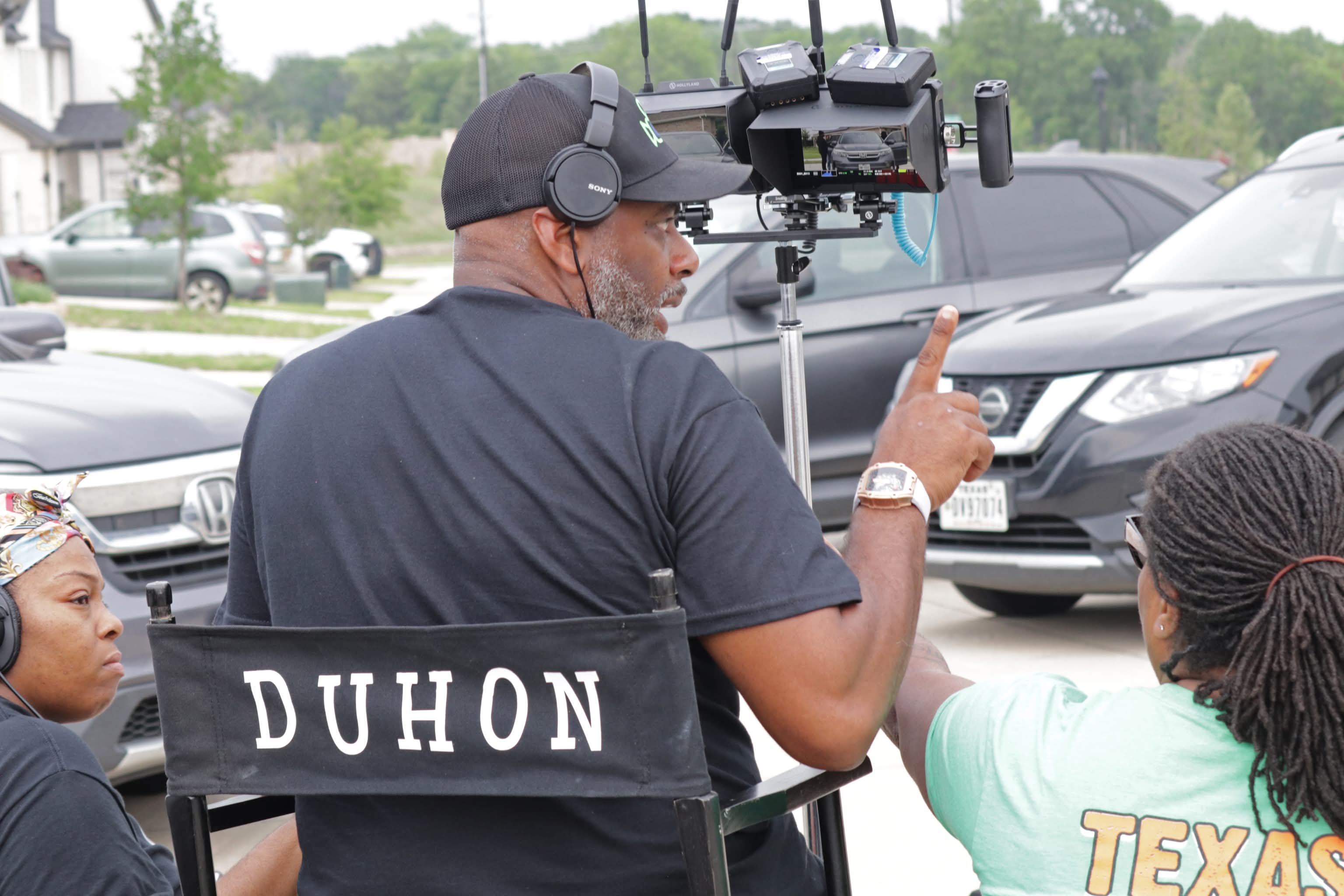 Man with headphones filming with a camera on a tripod, two women nearby, parking lot background