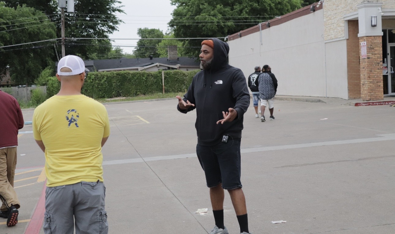 Two men talking outdoors on a street, one wearing a yellow shirt and white cap, the other in black hoodie and shorts.