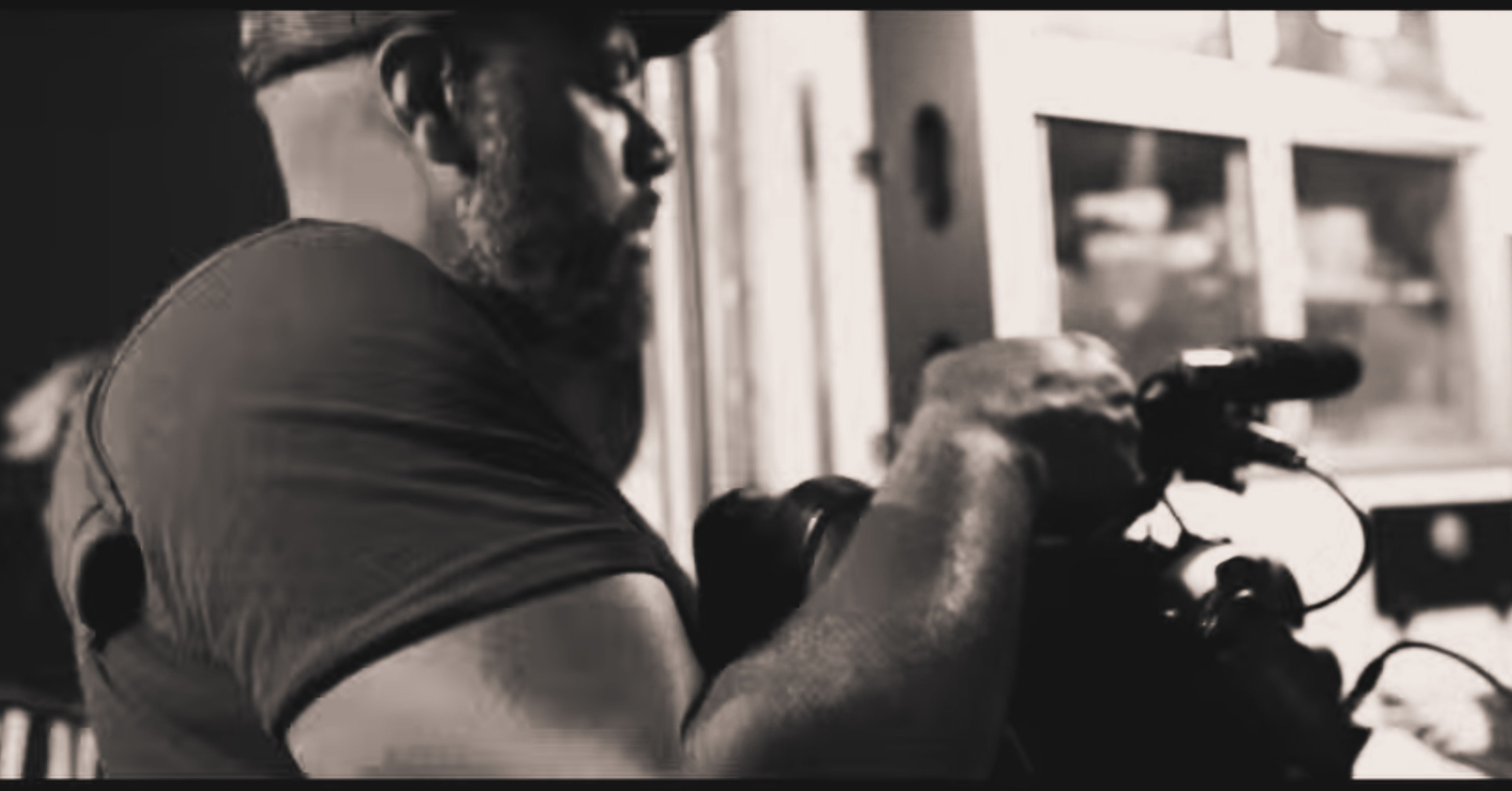 Man with a beard and glasses holding a camera, focusing on filming or taking a photo, in black and white.