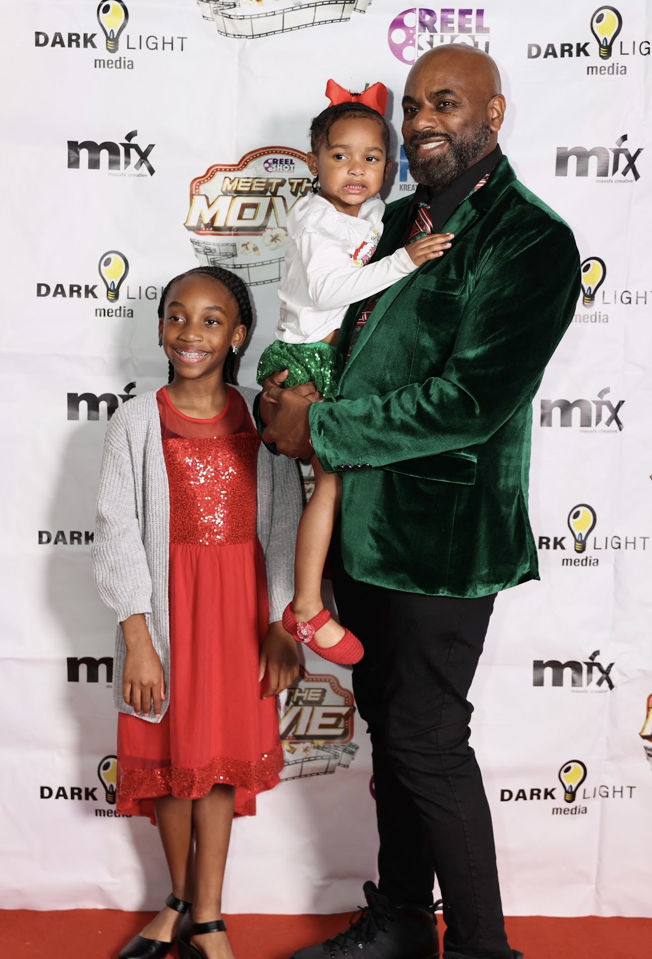 Man holding a young girl, girl standing beside a woman, all smiling, red and green outfits, backdrop with logos.