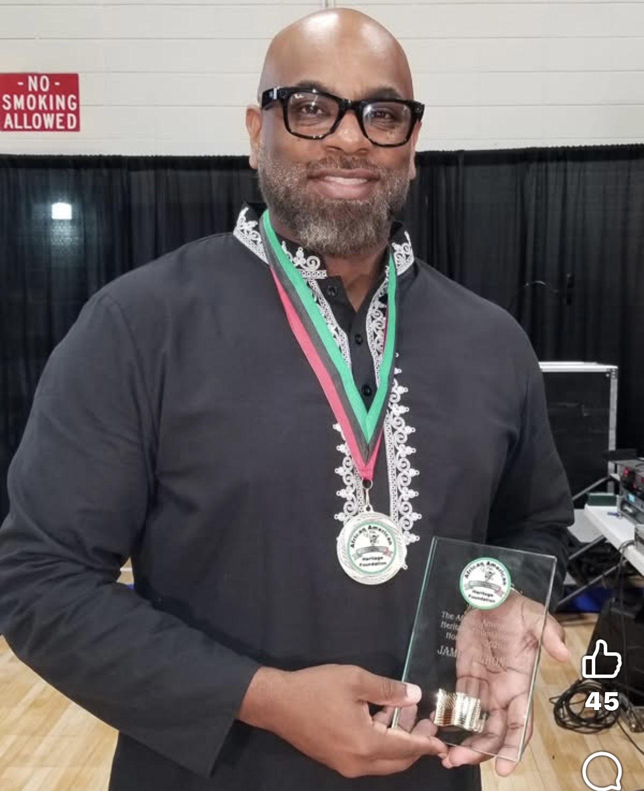 Man with glasses and beard holding a glass award, wearing medals around neck, smiling indoors.