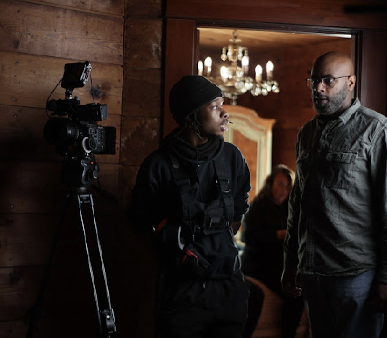 Two people talking indoors near a camera, with a chandelier and a woman in the background.