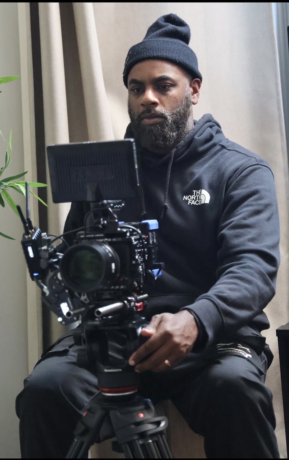Man with a beard wearing a beanie and hoodie, sitting behind a camera on a tripod, indoors.