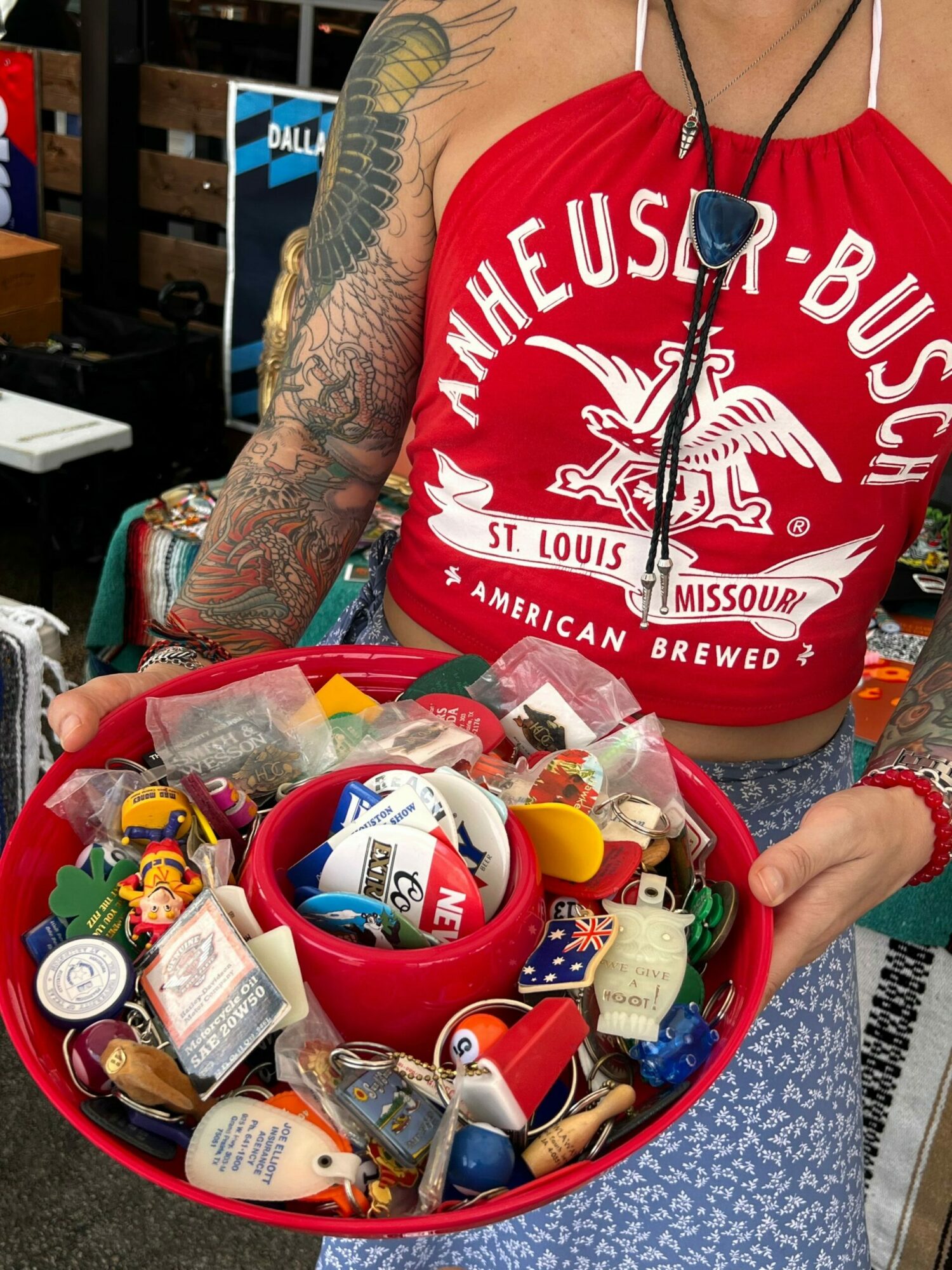 Person wearing a red T-shirt with white text and a tattooed arm, holding a red container filled with various small items.