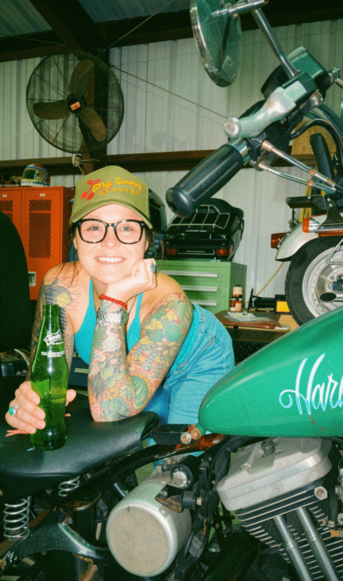 Smiling woman with glasses and tattoos sitting on a motorcycle in a garage, holding a green bottle.