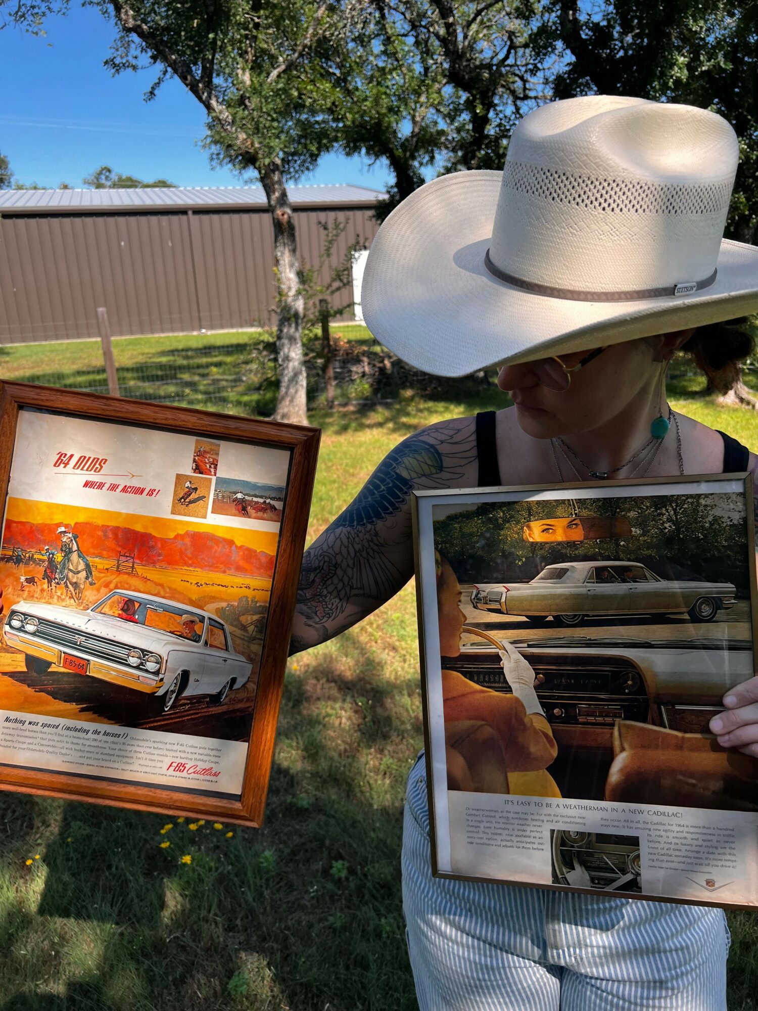 A white hat and a mirror reflecting a person outdoors with trees and a fence, a framed poster of a car, and a wooden framed picture of a sunset and a car.