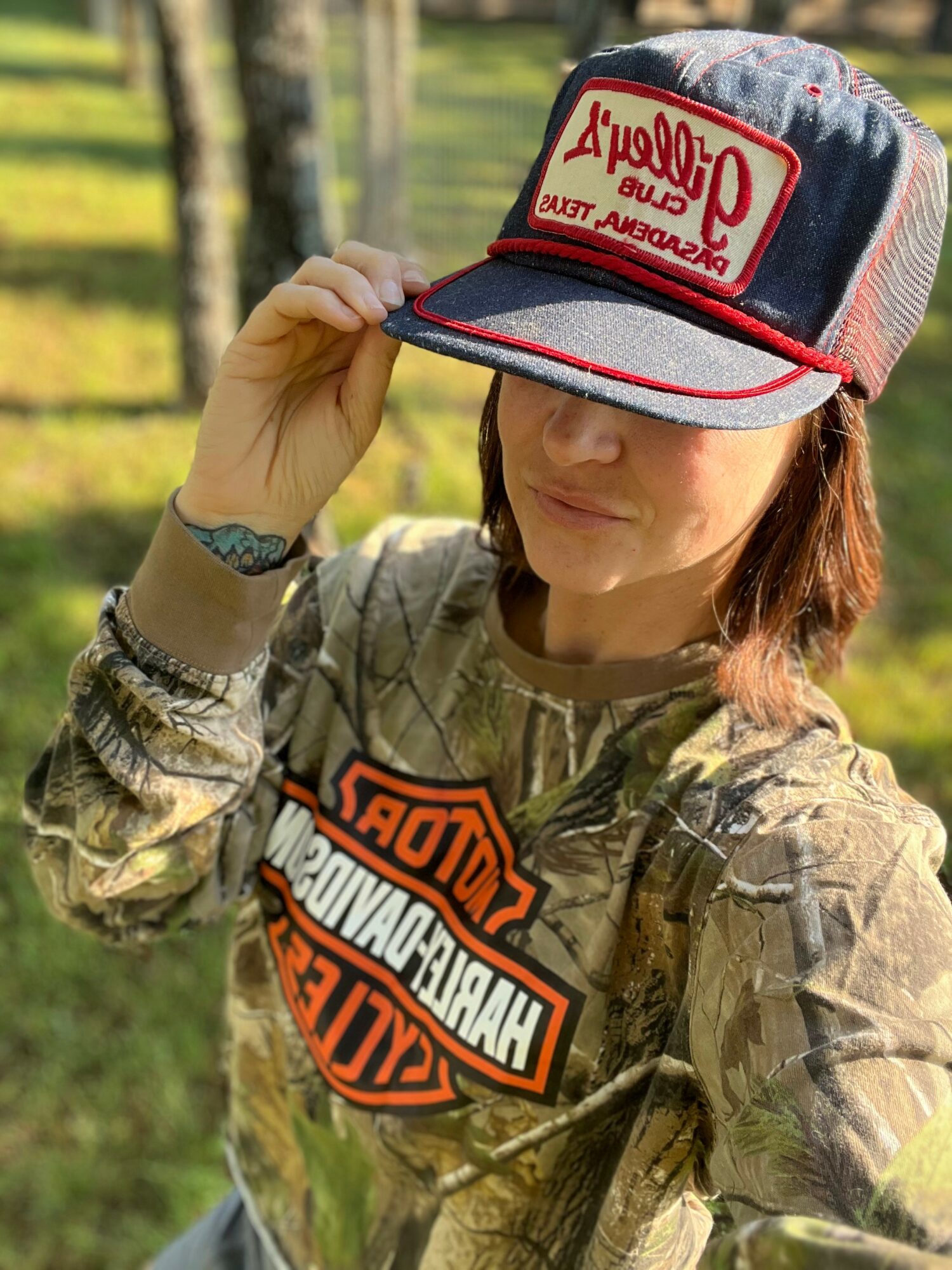 Woman outdoors wearing a camouflage jacket and a Harley-Davidson patch, holding a cap with a red and white logo, in a grassy area with trees.