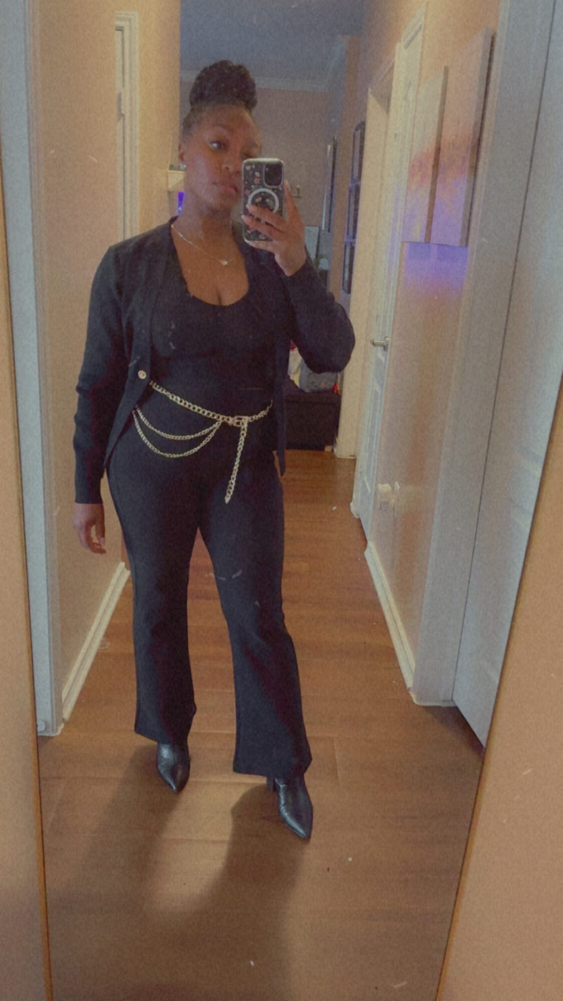 Person taking a mirror selfie in a hallway, wearing dark clothing and a chain belt, with a wooden floor and light-colored walls.