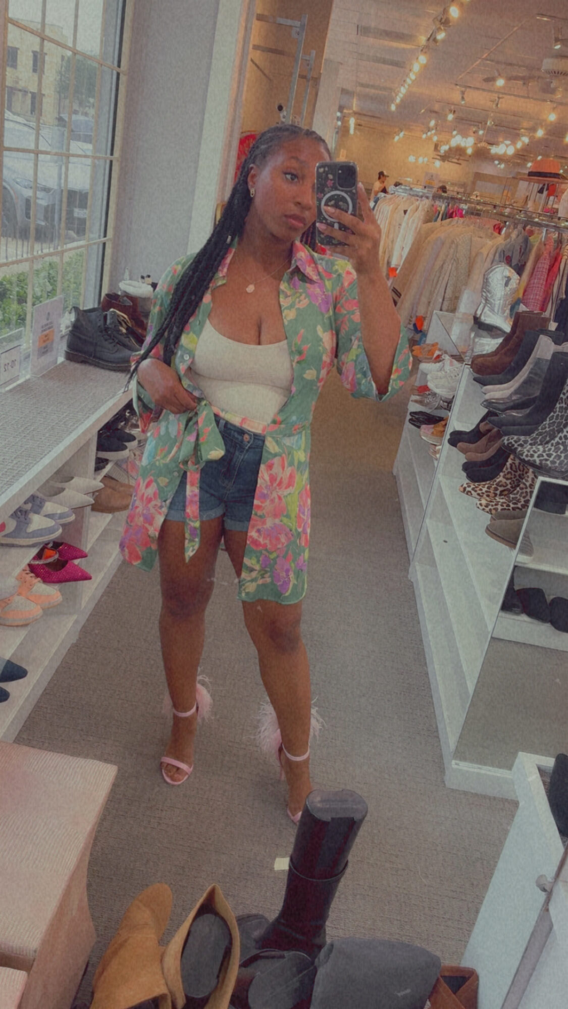 Person taking mirror selfie in clothing store, wearing a colorful jacket, white top, and high heels.