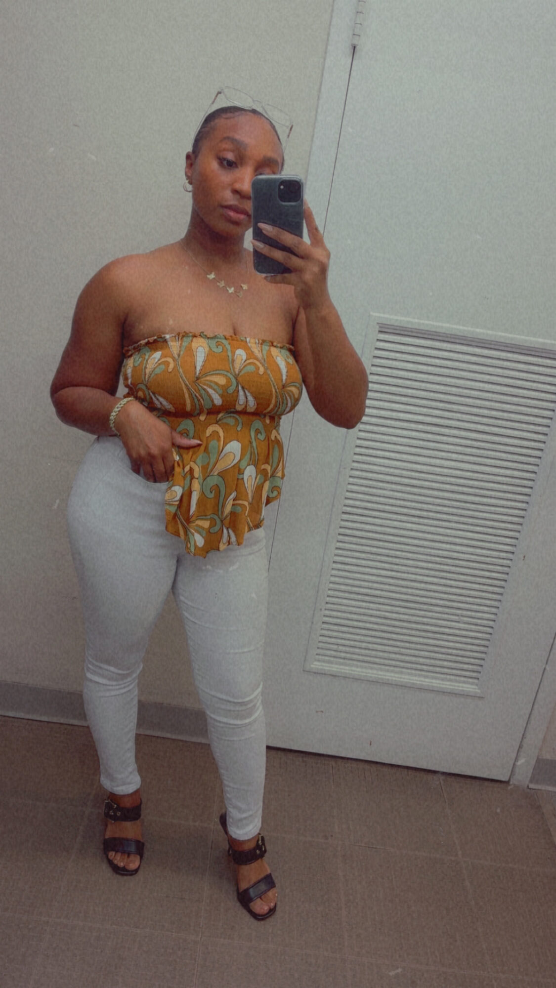 Woman taking mirror selfie, wearing a strapless top with a yellow and white pattern, white pants, and black sandals.