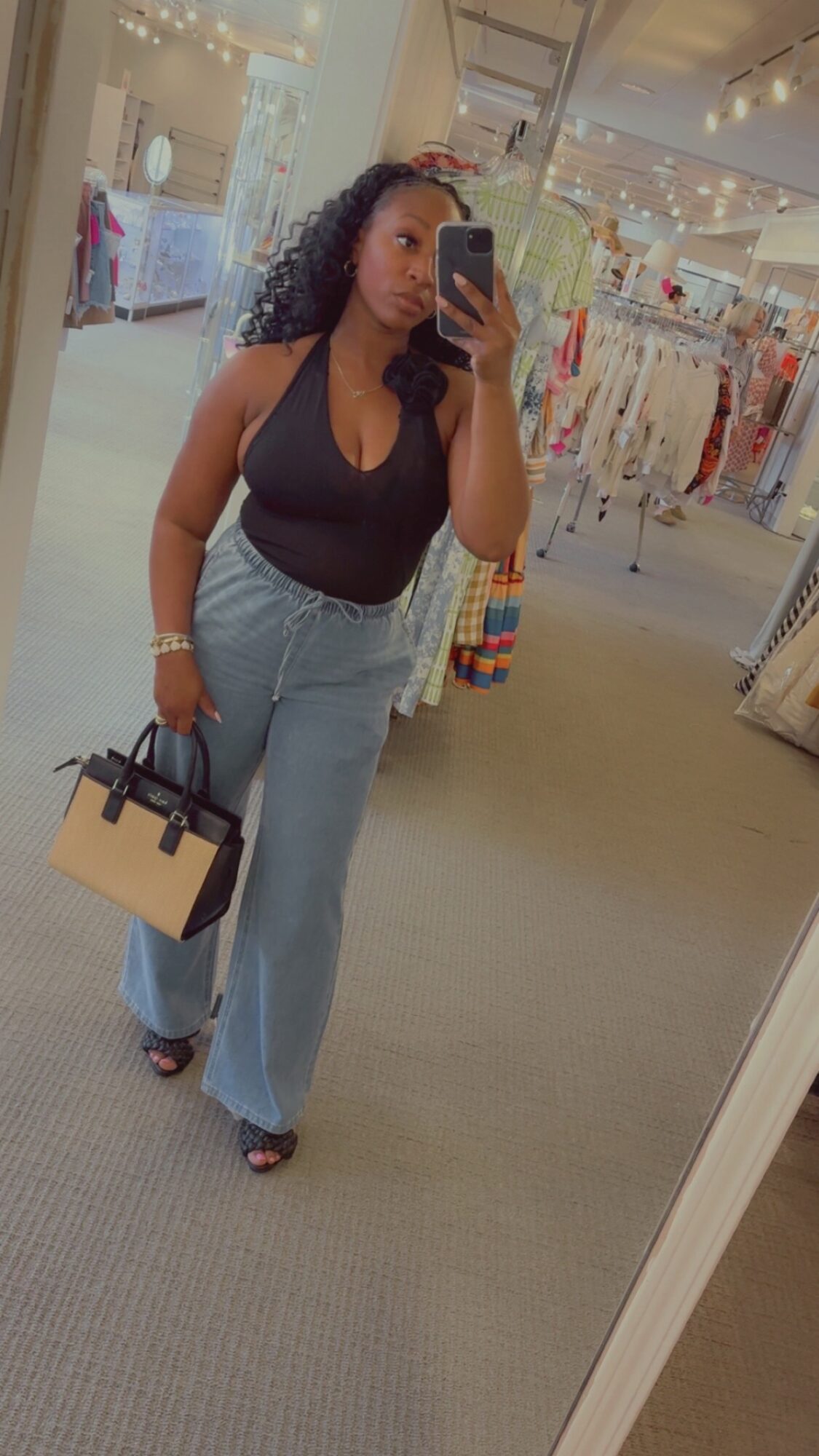 Woman taking mirror selfie in clothing store, holding a handbag, wearing a black top and light blue jeans.