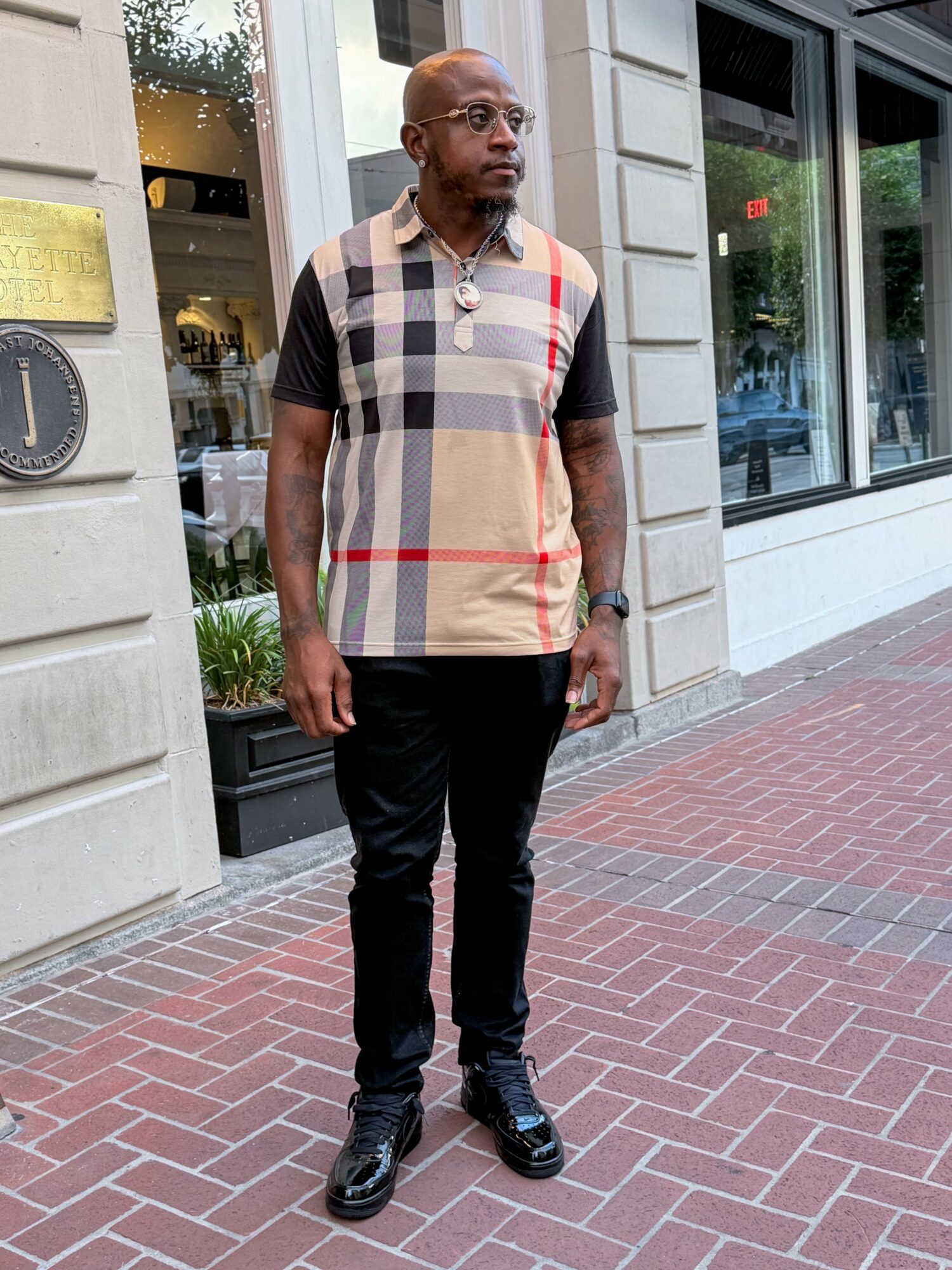 Man standing on sidewalk outside building, wearing glasses, a beige plaid shirt with red and black accents, black pants, and sneakers.