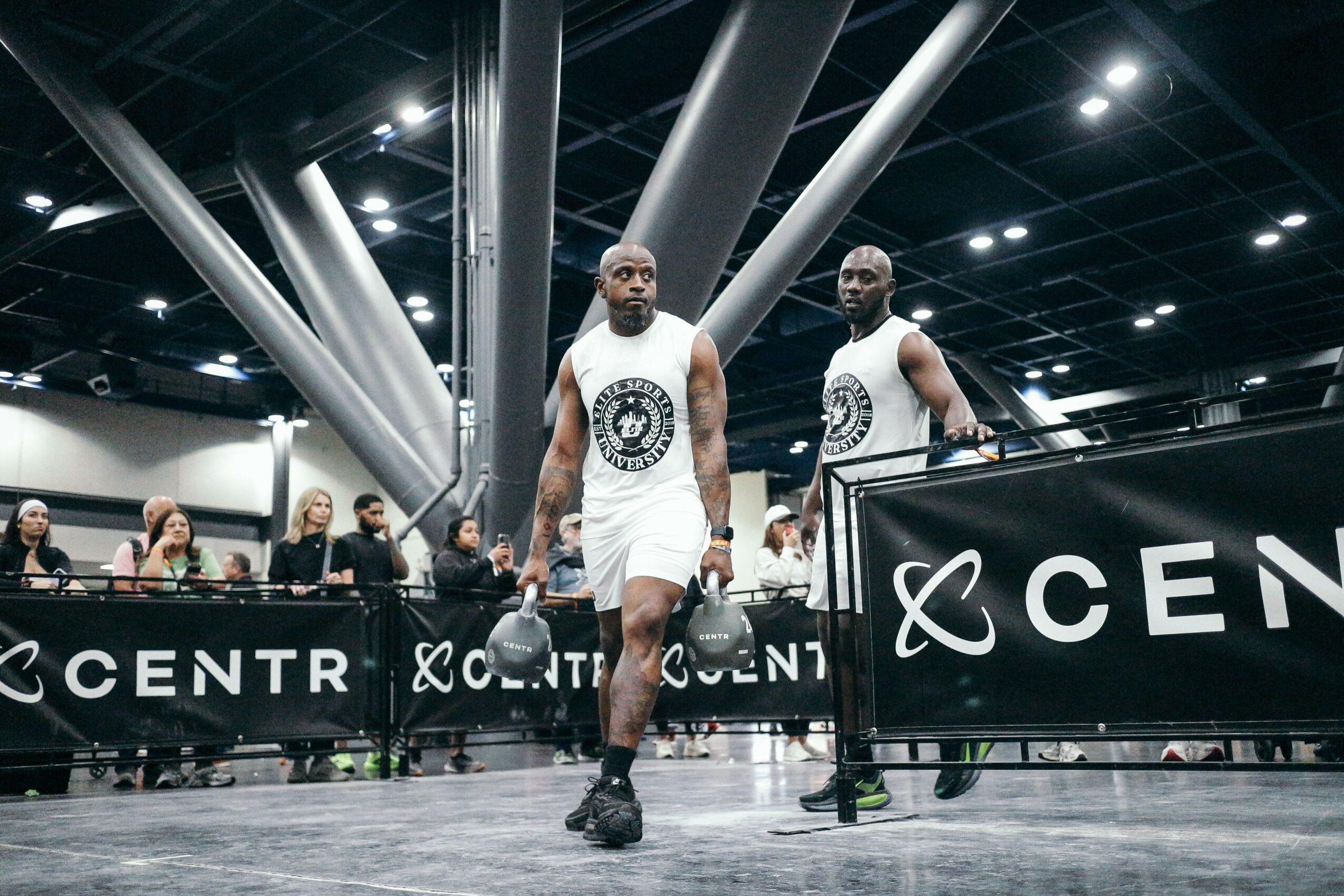 Two basketball players in white uniforms walk on a court with a black barrier and audience in the background.