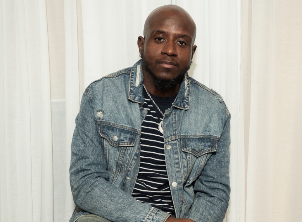 Man with a shaved head and beard wearing a denim jacket and striped shirt, sitting in front of white curtains.