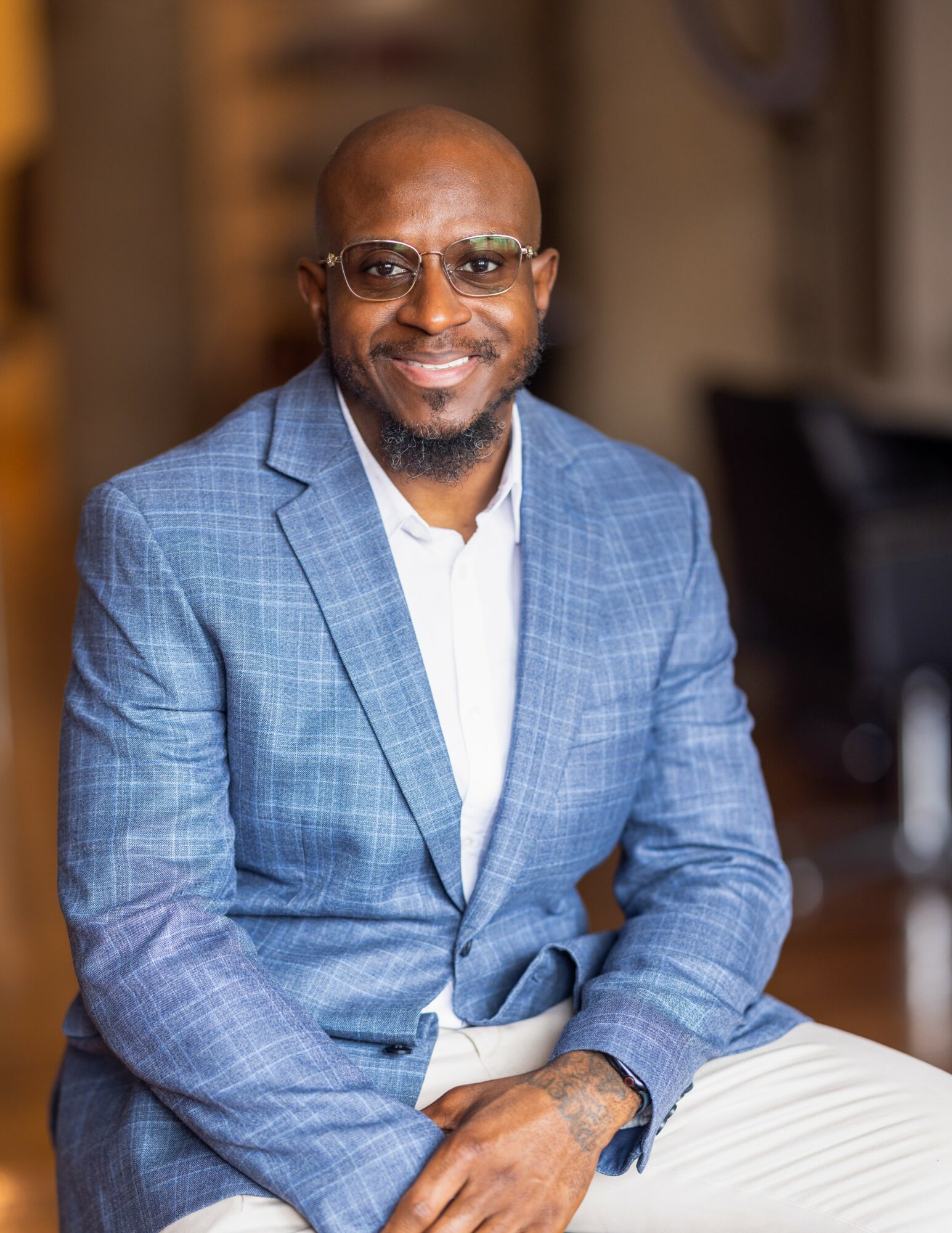 Man in blue checkered suit with glasses, smiling, sitting indoors with blurred background.