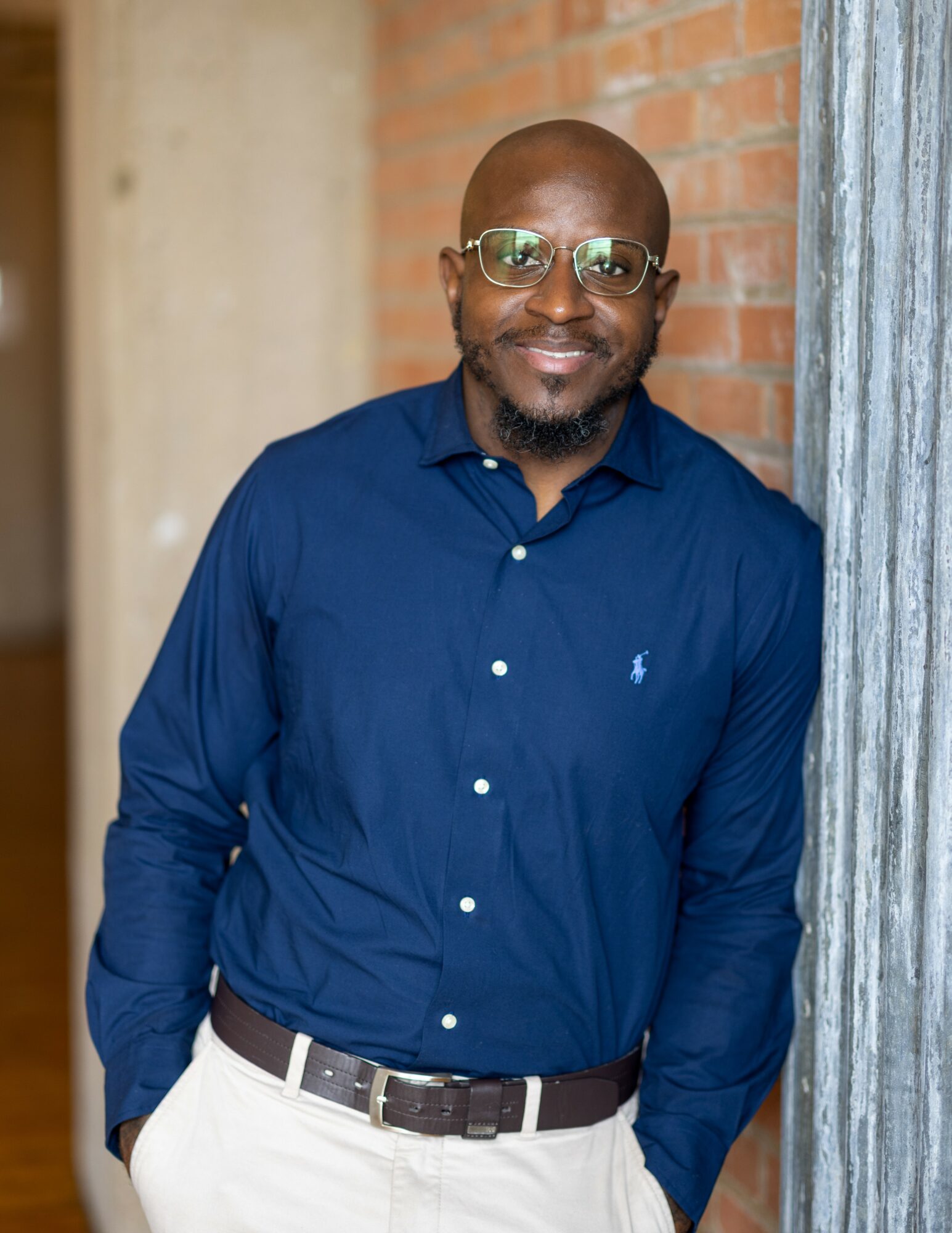 Man with glasses and a beard wearing a navy blue shirt and white pants, standing outdoors near a brick wall.