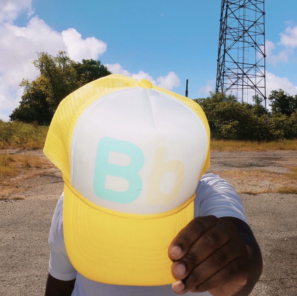 Person holding a yellow and white cap with a logo, outdoors with trees and a tower in background.