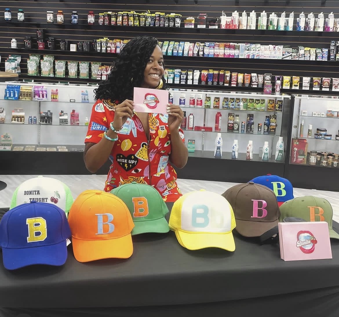 Woman smiling behind a table with colorful caps and a card in a store aisle, shelves with products in background.