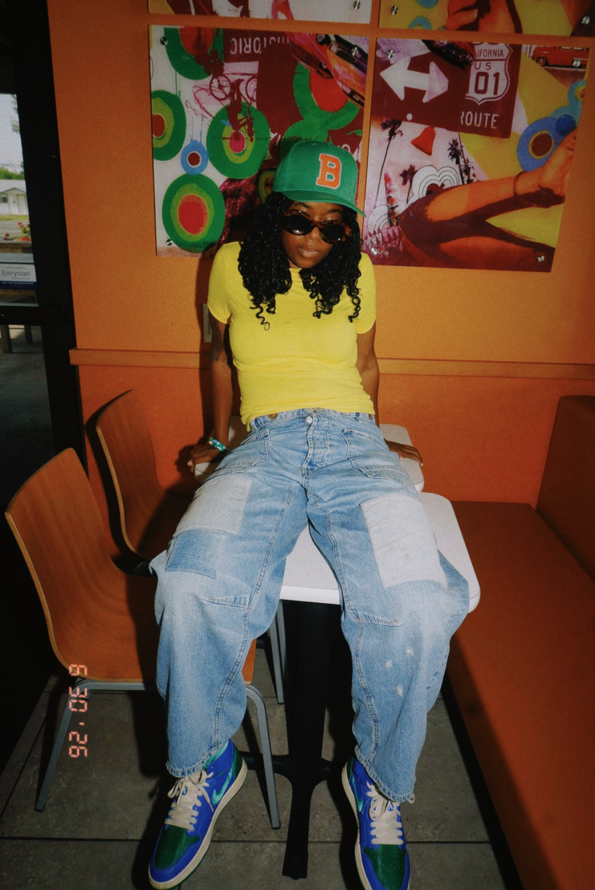 Person with curly hair wearing sunglasses, a green cap, yellow top, and baggy jeans sitting in a colorful indoor setting.