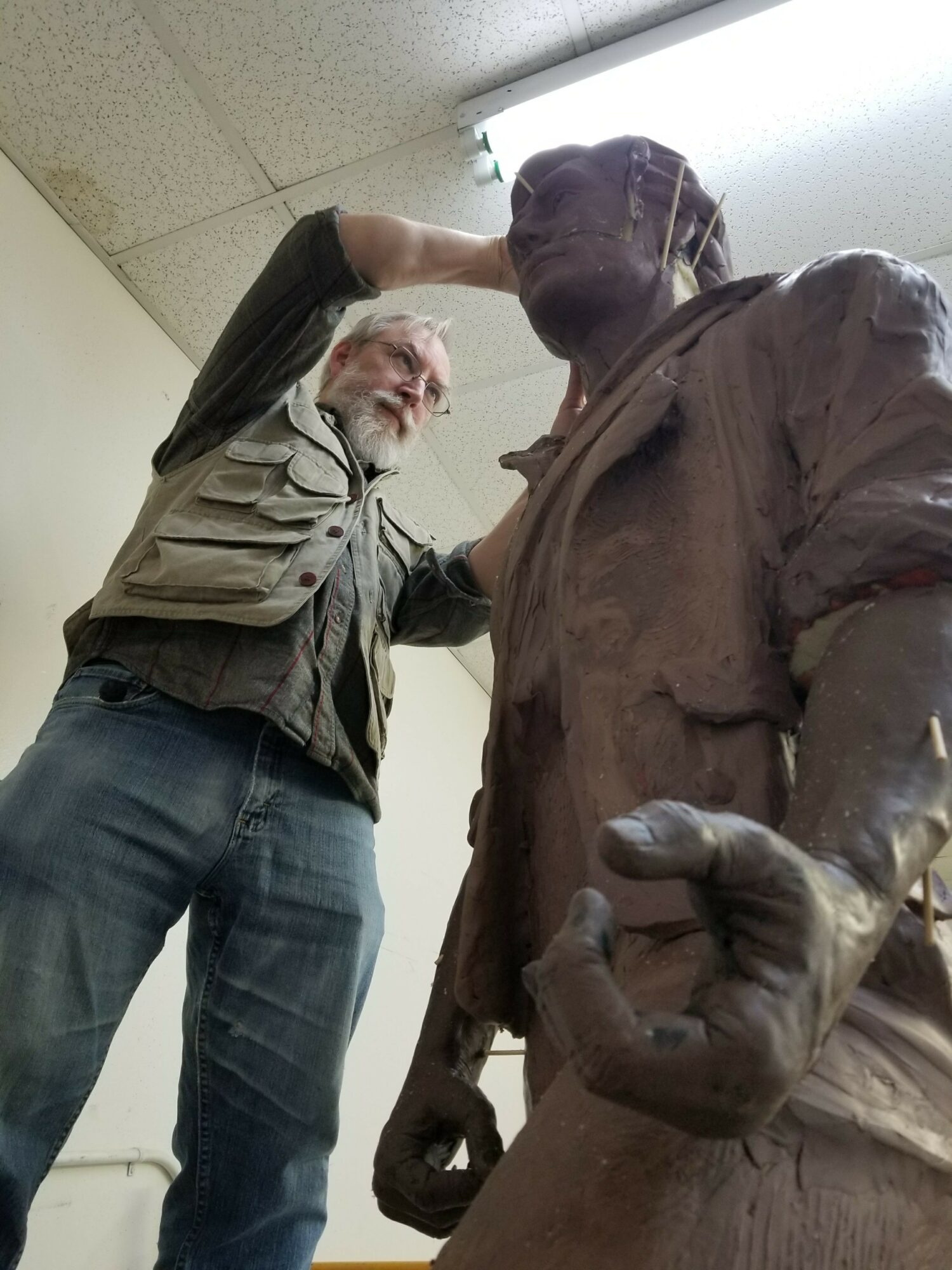 Man working on a large bronze statue in an indoor setting, ceiling light visible above.