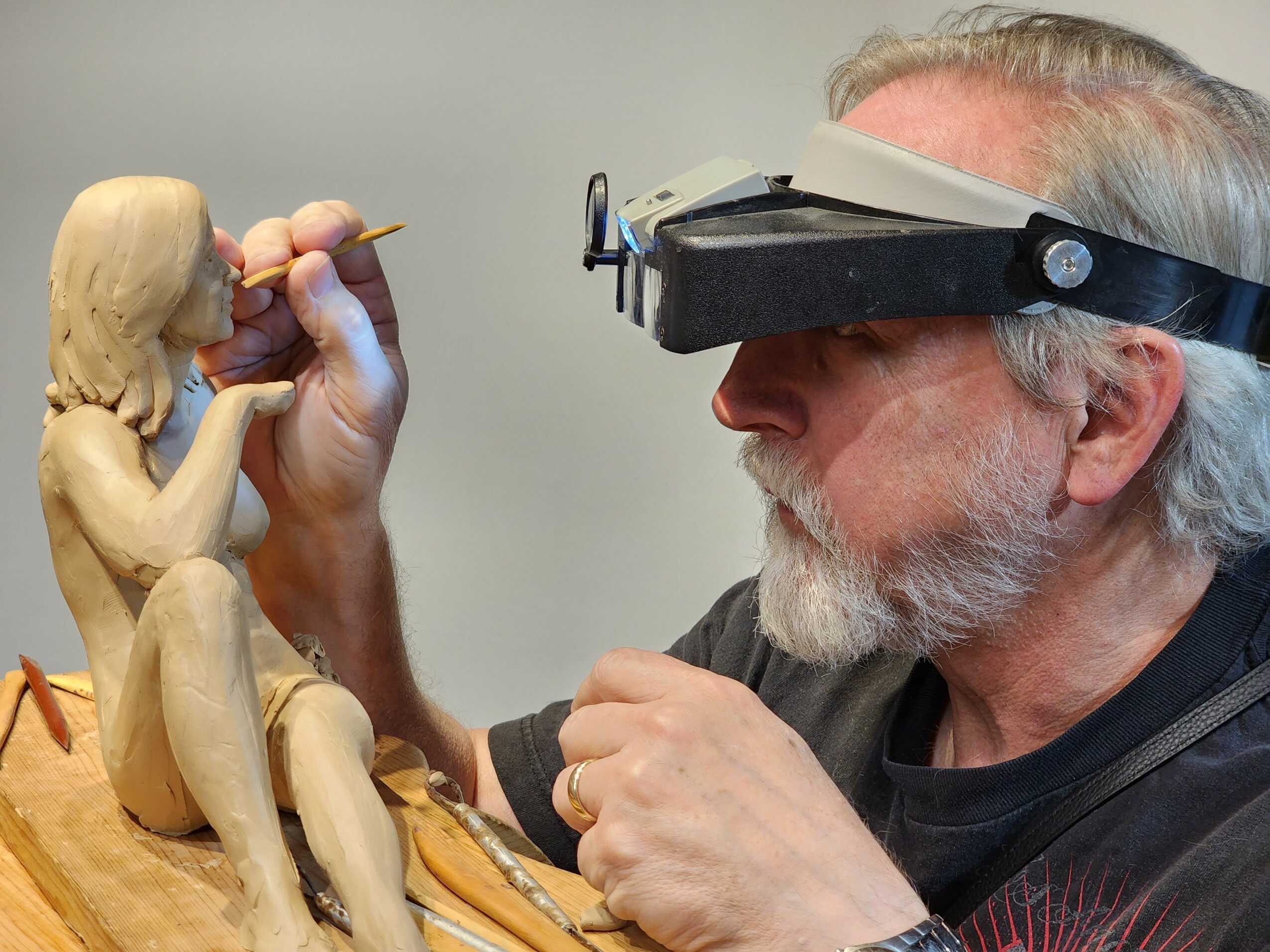 Artist carves a wooden sculpture of a seated woman with long hair, using a small tool, while wearing magnifying glasses.