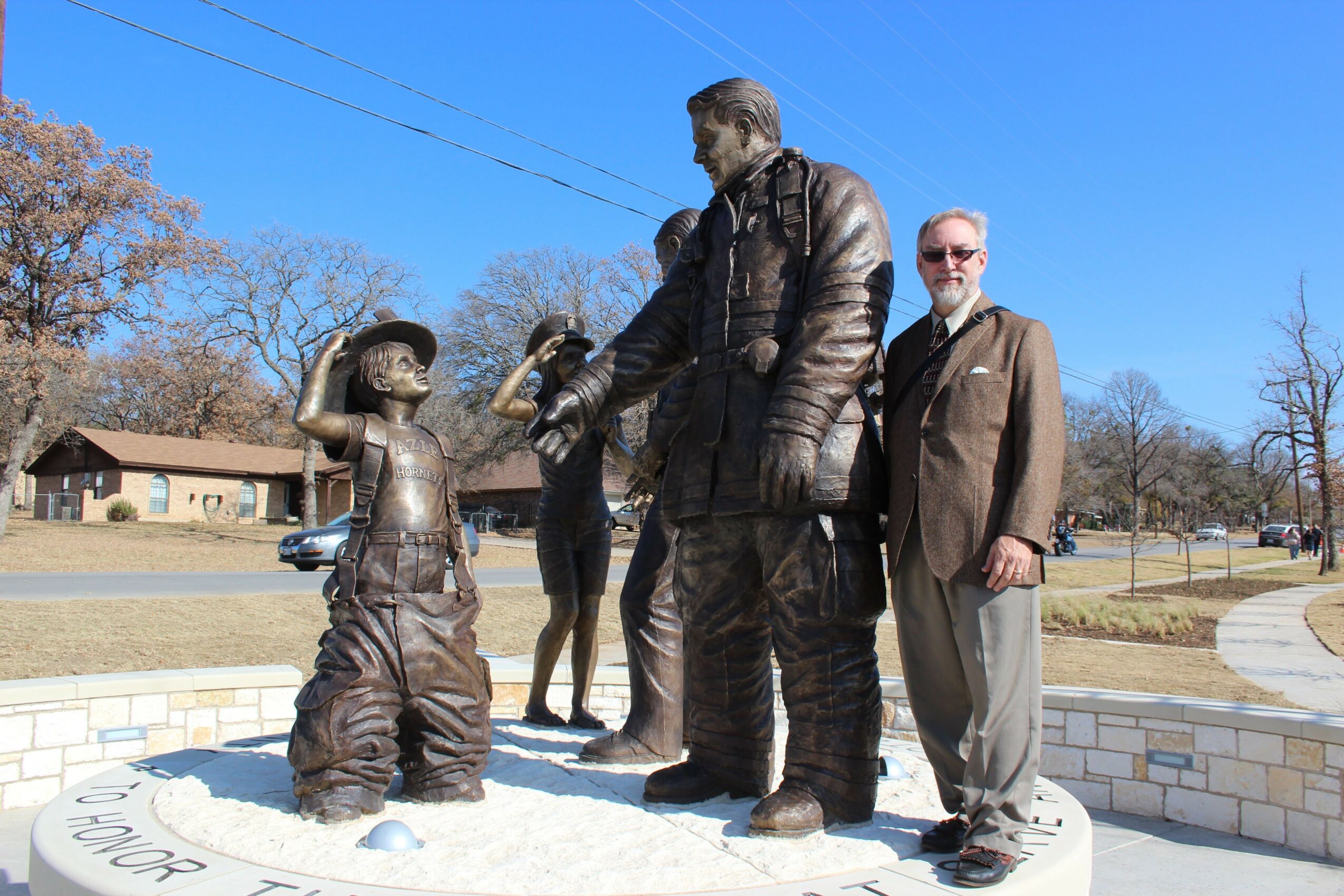 Man standing next to a bronze statue of a boy saluting a soldier, in a park with trees and buildings in the background.