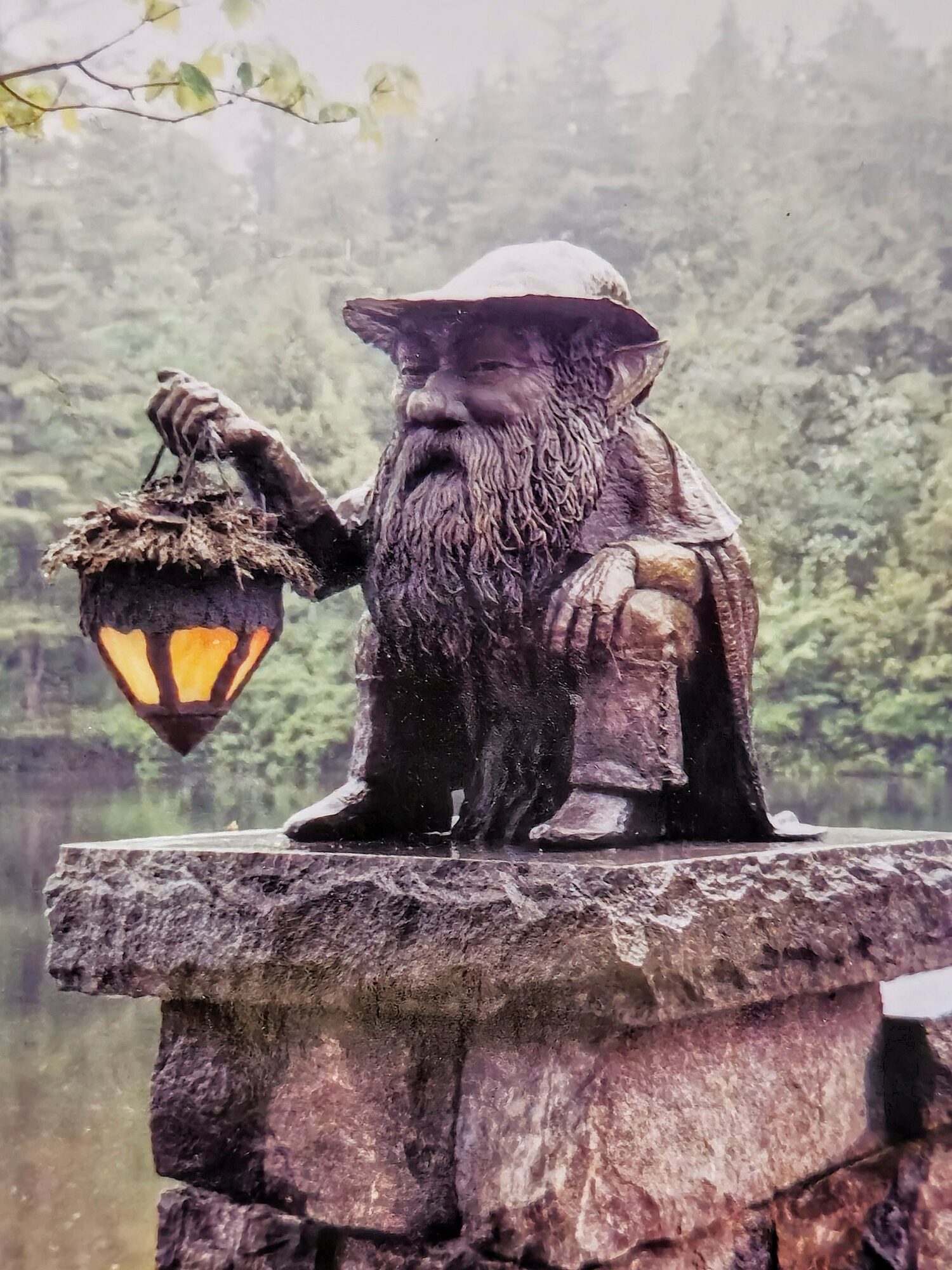 Gnome statue with a beard, hat, and lantern, sitting on a stone platform outdoors with trees in the background.