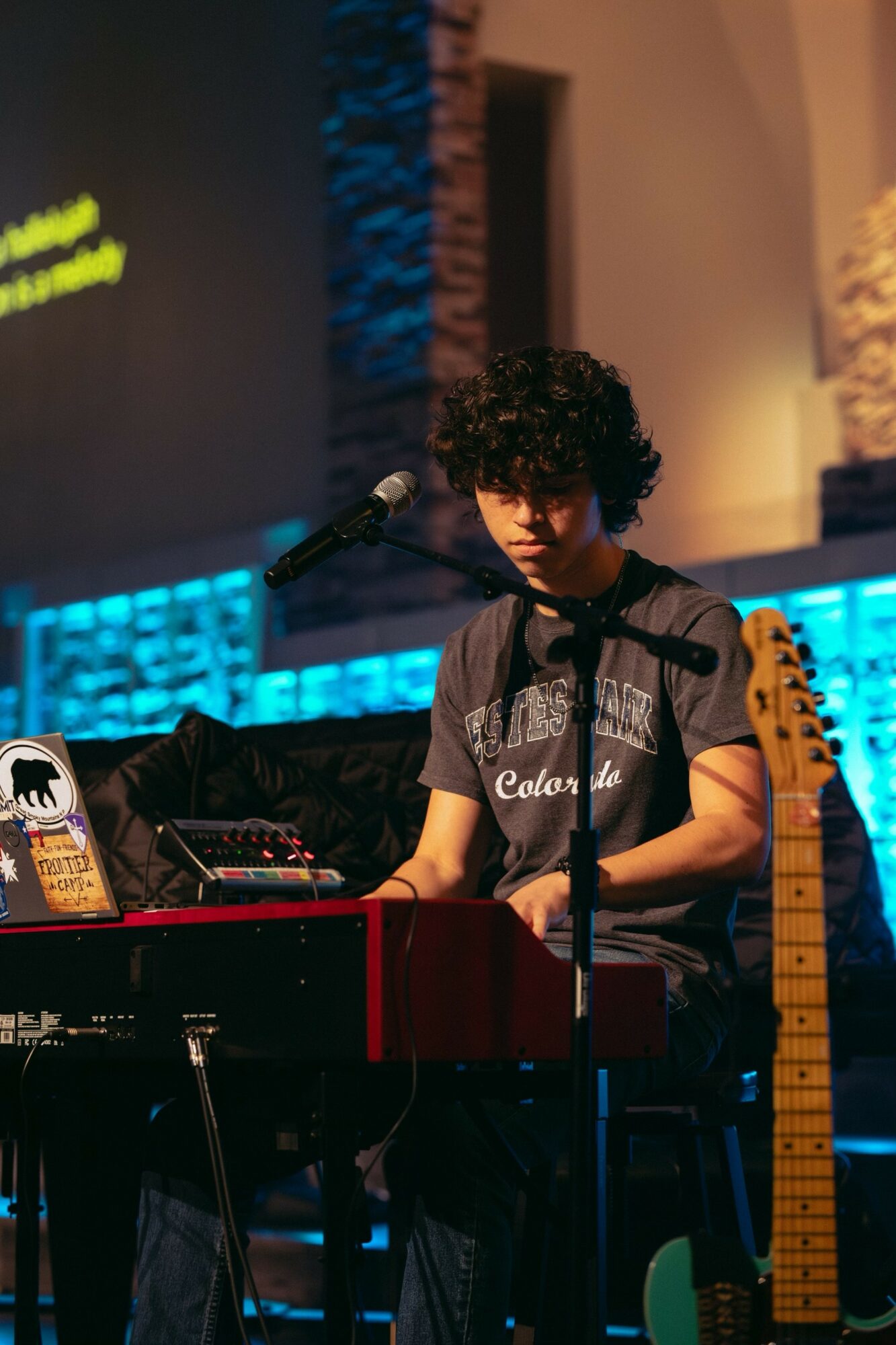 Young person with curly hair playing keyboard on stage with microphone, illuminated by colorful stage lights.
