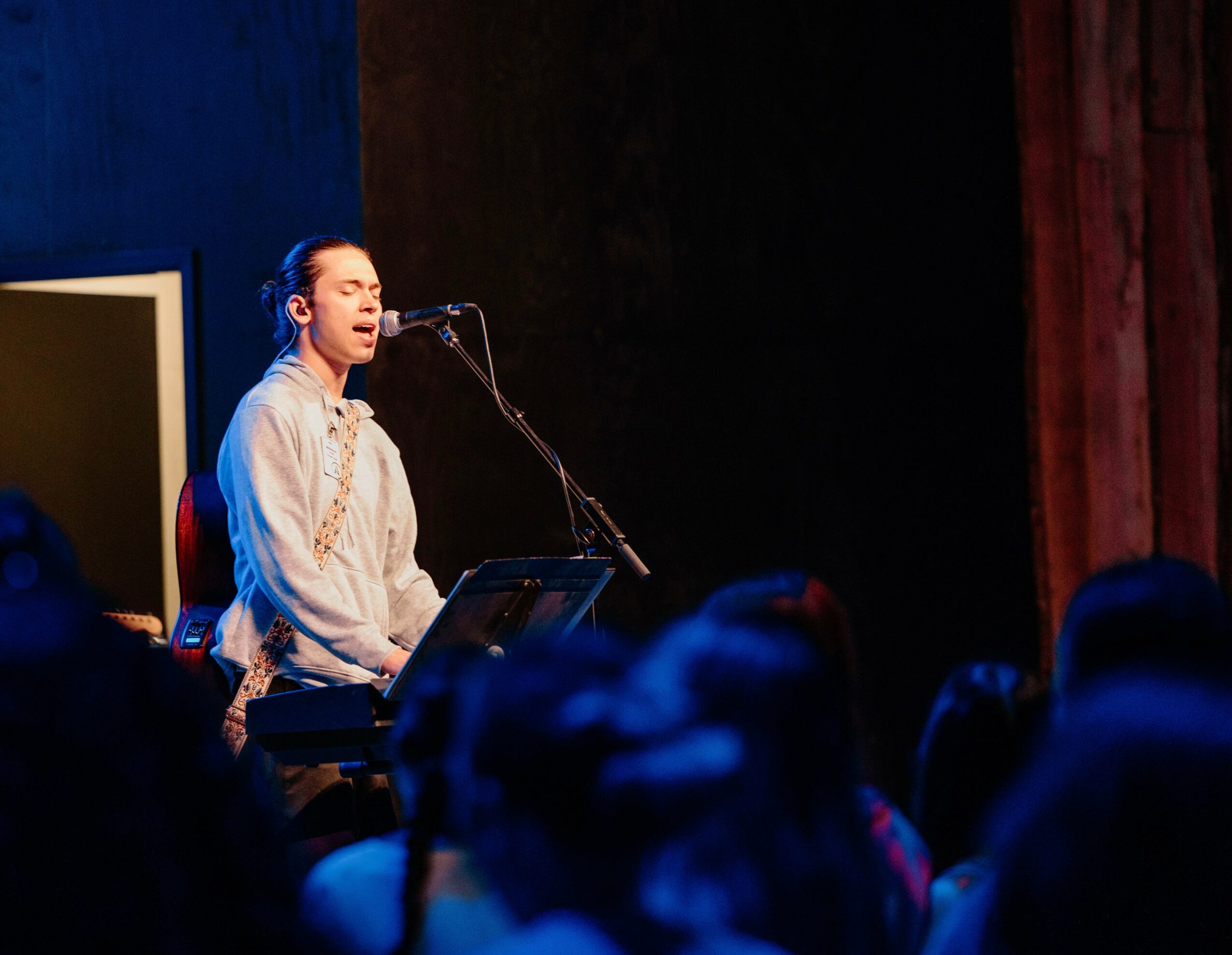 Person singing and playing keyboard on stage with audience in foreground, dark background, stage lighting.