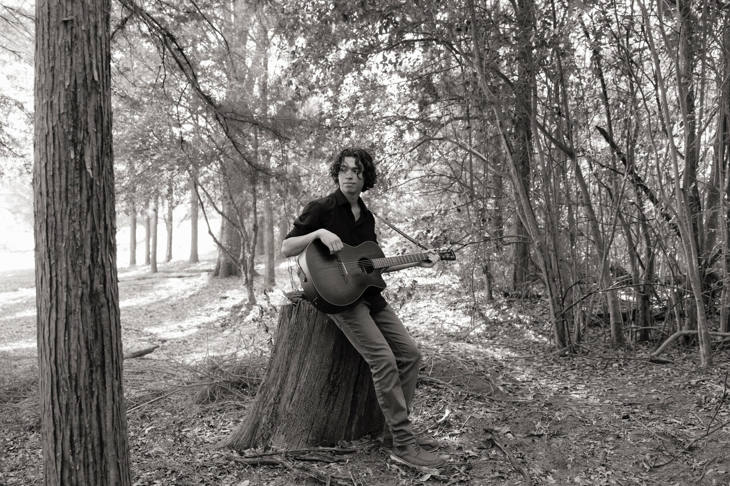 Person with curly hair playing guitar outdoors, leaning against a tree trunk in a wooded area.