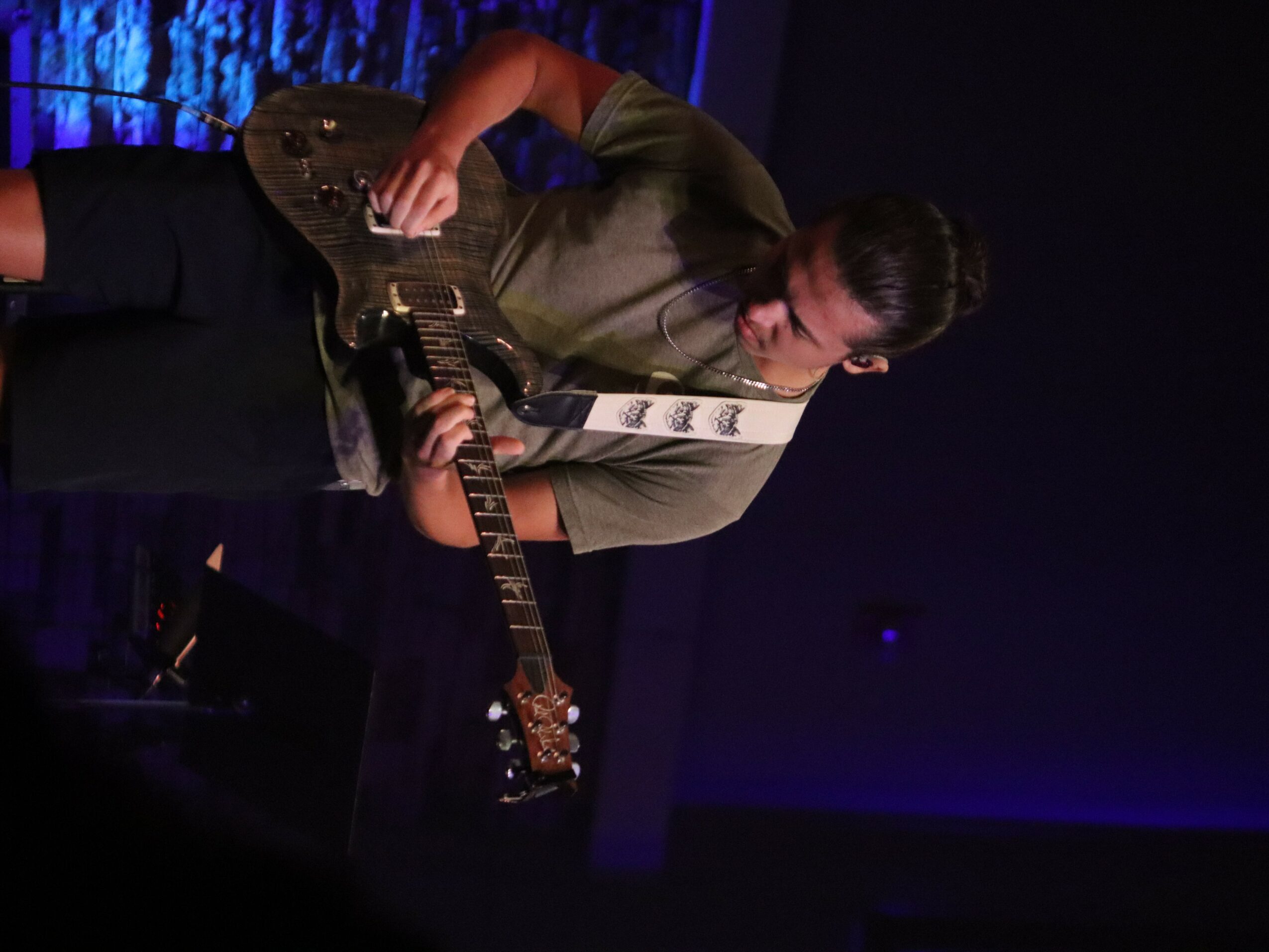 Person playing an electric guitar on stage with dark background and blue lighting, focused on the instrument.