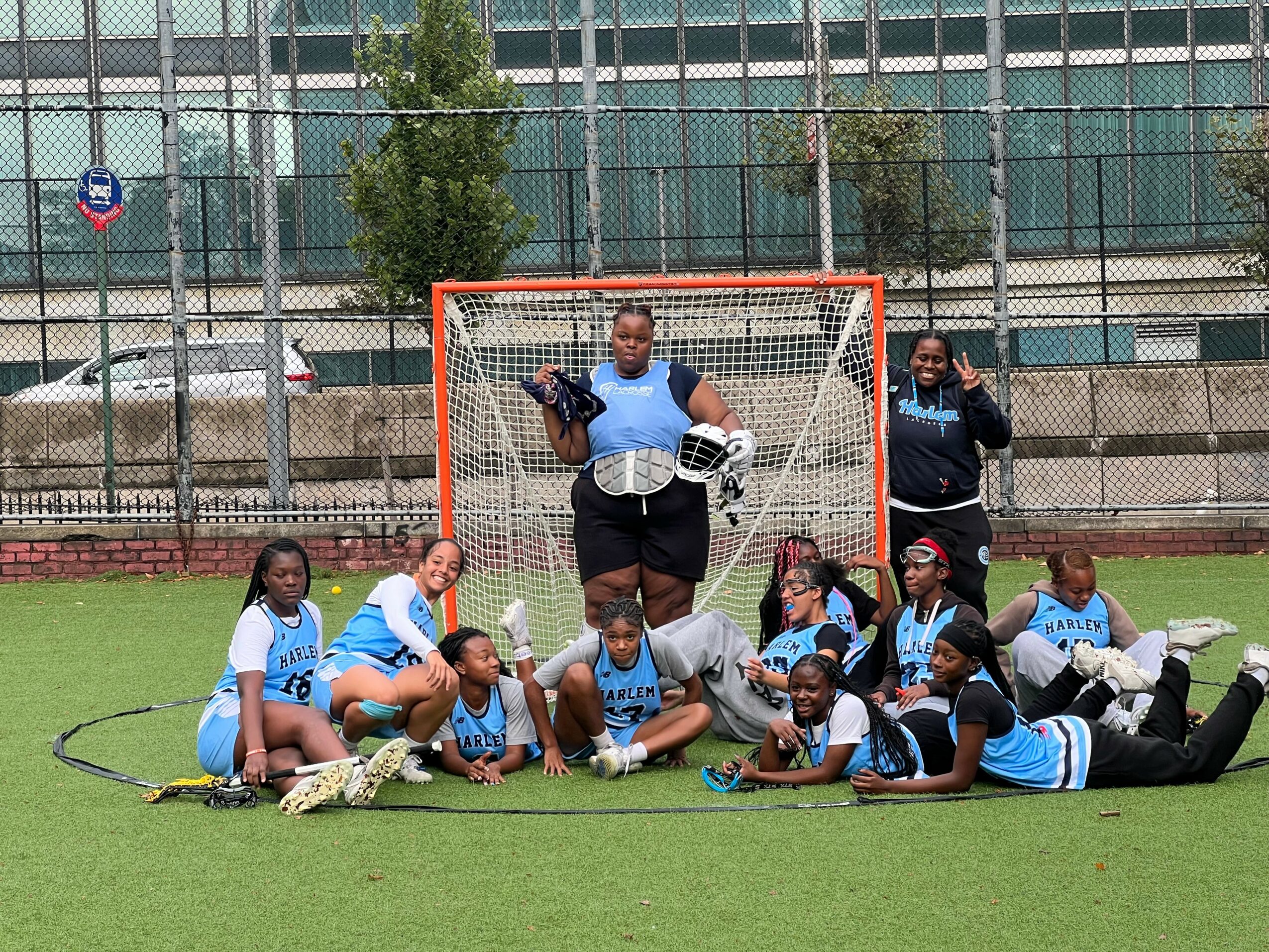 Group of young female field hockey players lying and sitting on the grass in front of a goalpost, smiling and posing.