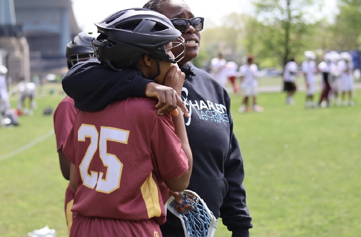 Two people hugging outdoors, one in a sports uniform with the number 25, the other in a dark hoodie, with a group in the background.