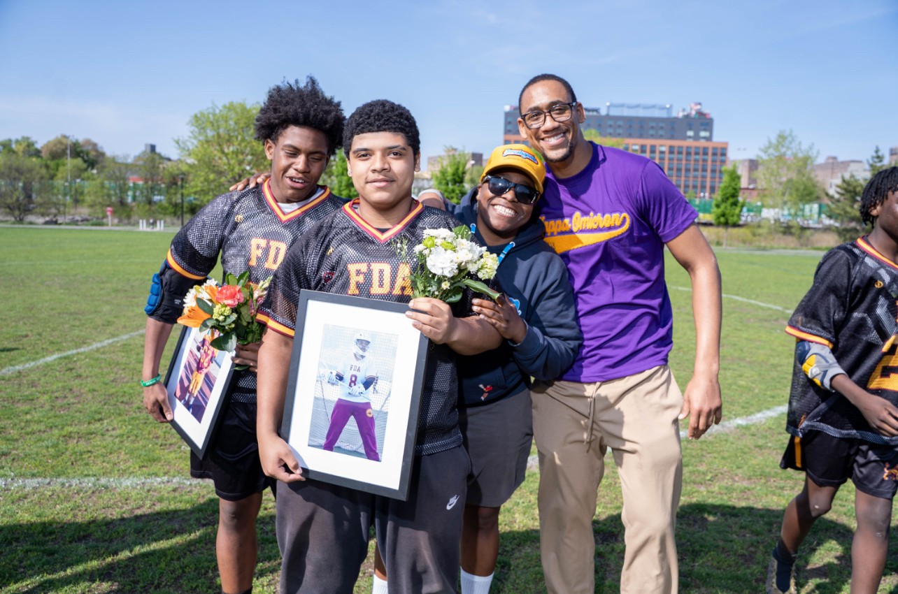 Group of four people outdoors on a grassy field, smiling, holding flowers and a framed picture, with trees and buildings in background.