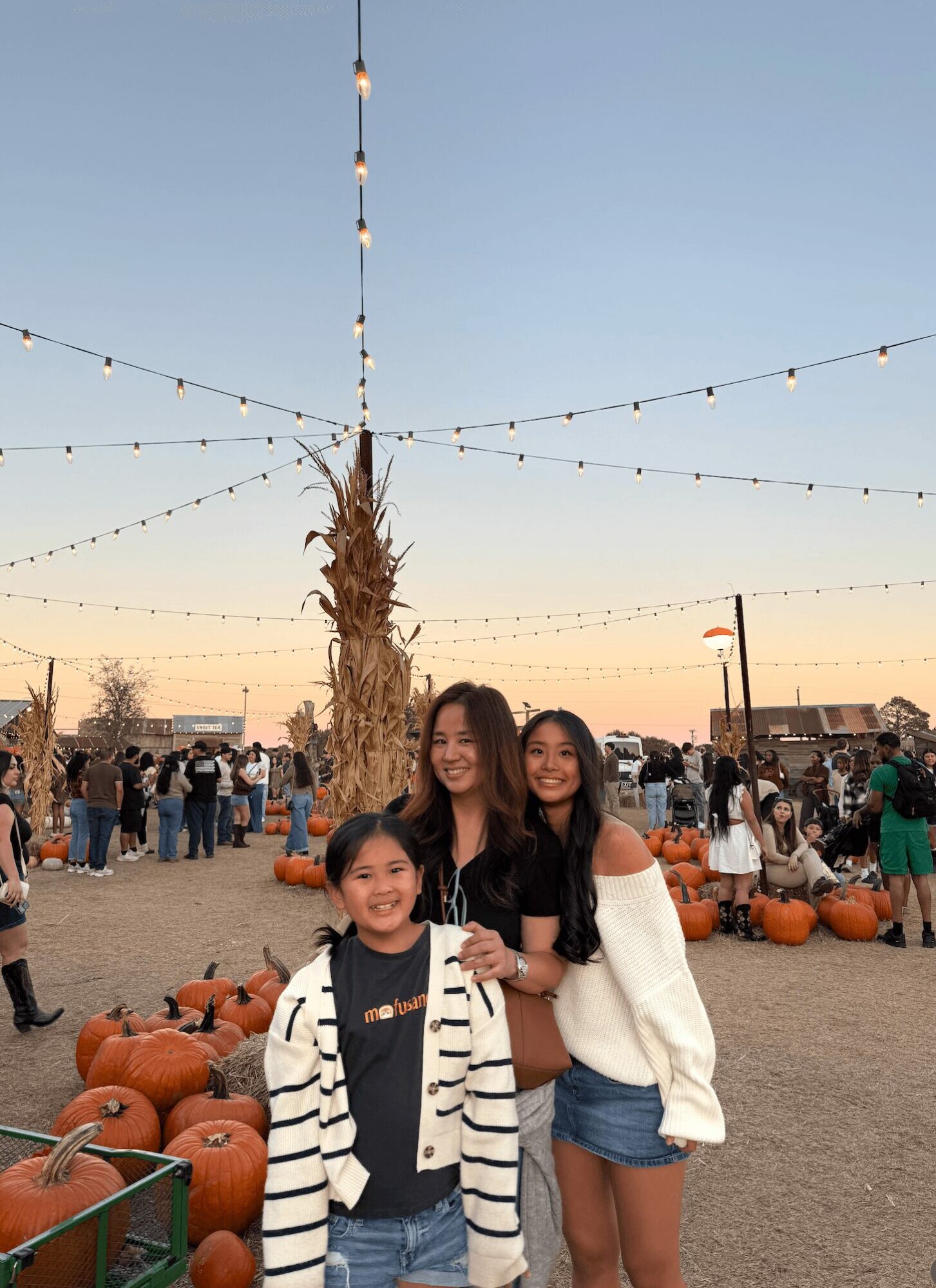 Three smiling girls standing outdoors at a pumpkin patch during sunset, with pumpkins and string lights in the background.