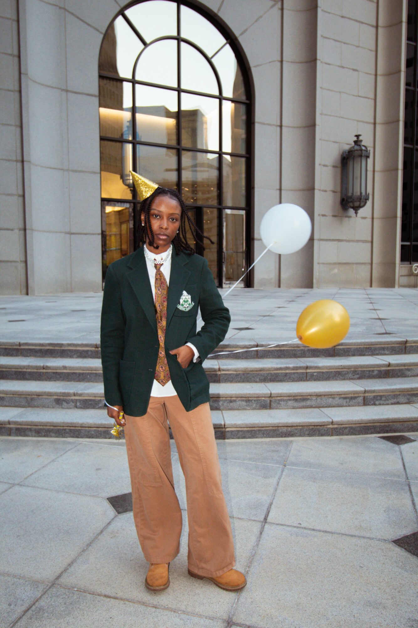 Person standing on steps in front of a building with large arched window, holding balloons, wearing a blazer and wide pants.