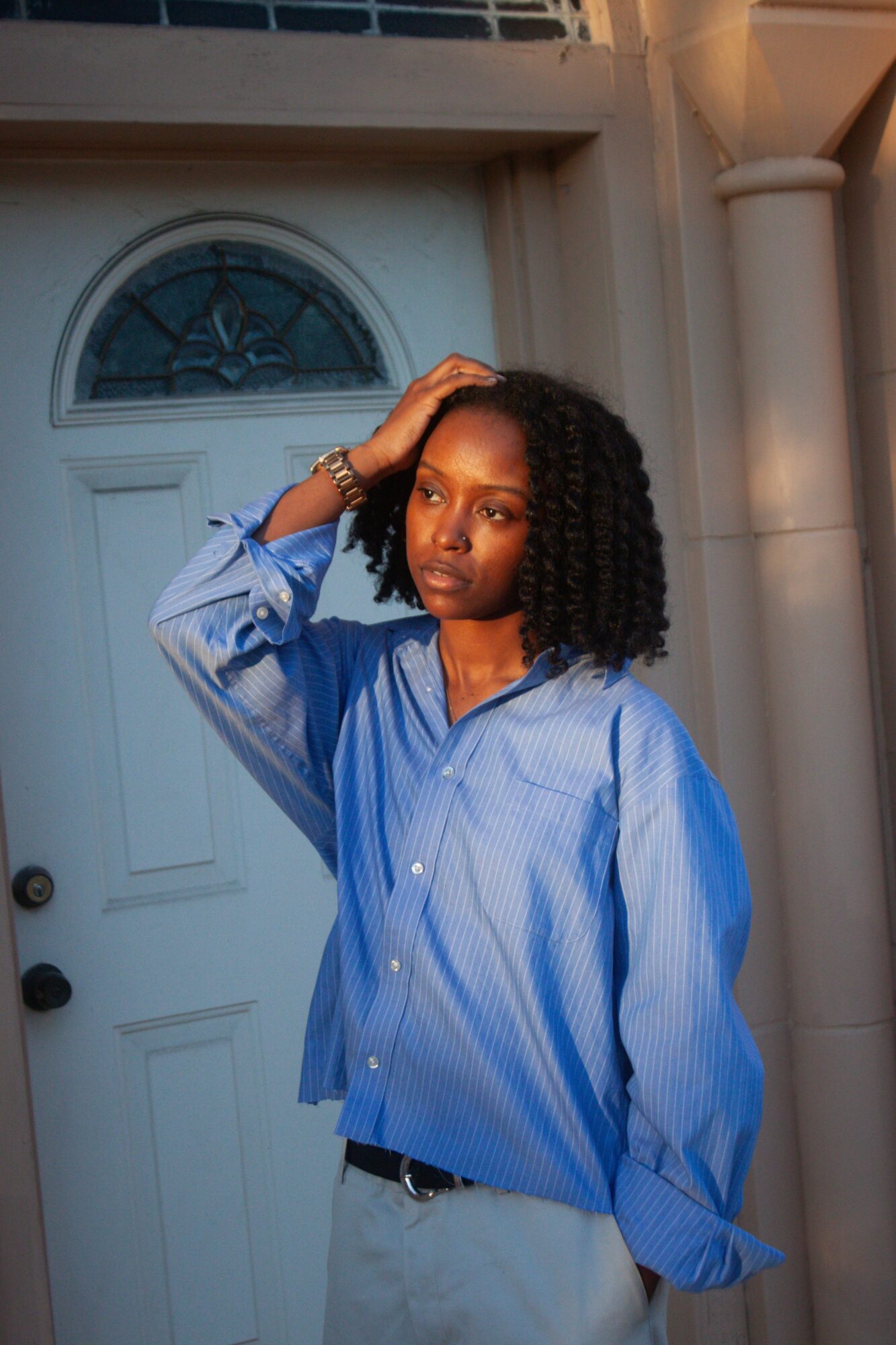 Young woman with curly hair wearing a blue striped shirt standing outside near a door, sunlight on her face.