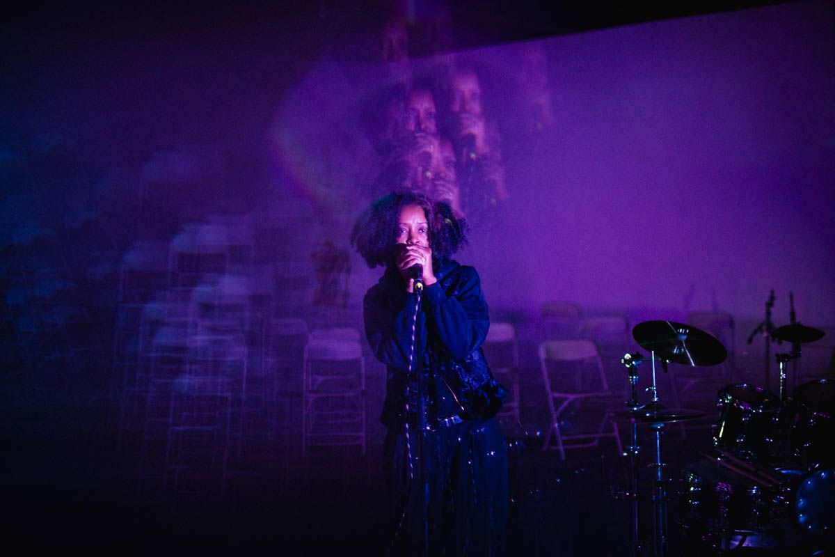 Person singing into a microphone on stage with purple lighting, musical instruments nearby, and chairs in the background.