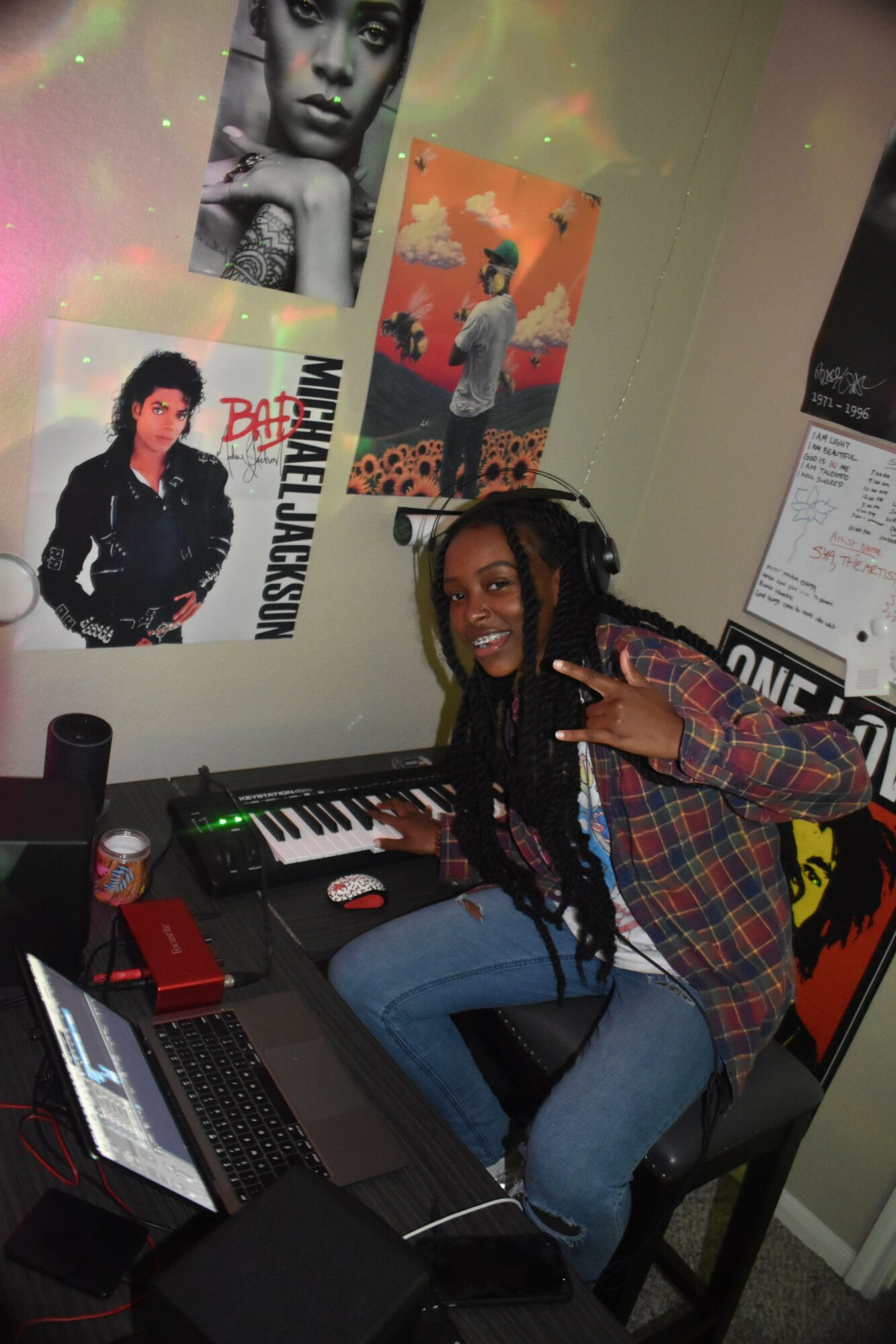 Person with long braided hair sitting at a desk with music equipment and posters on the wall behind.