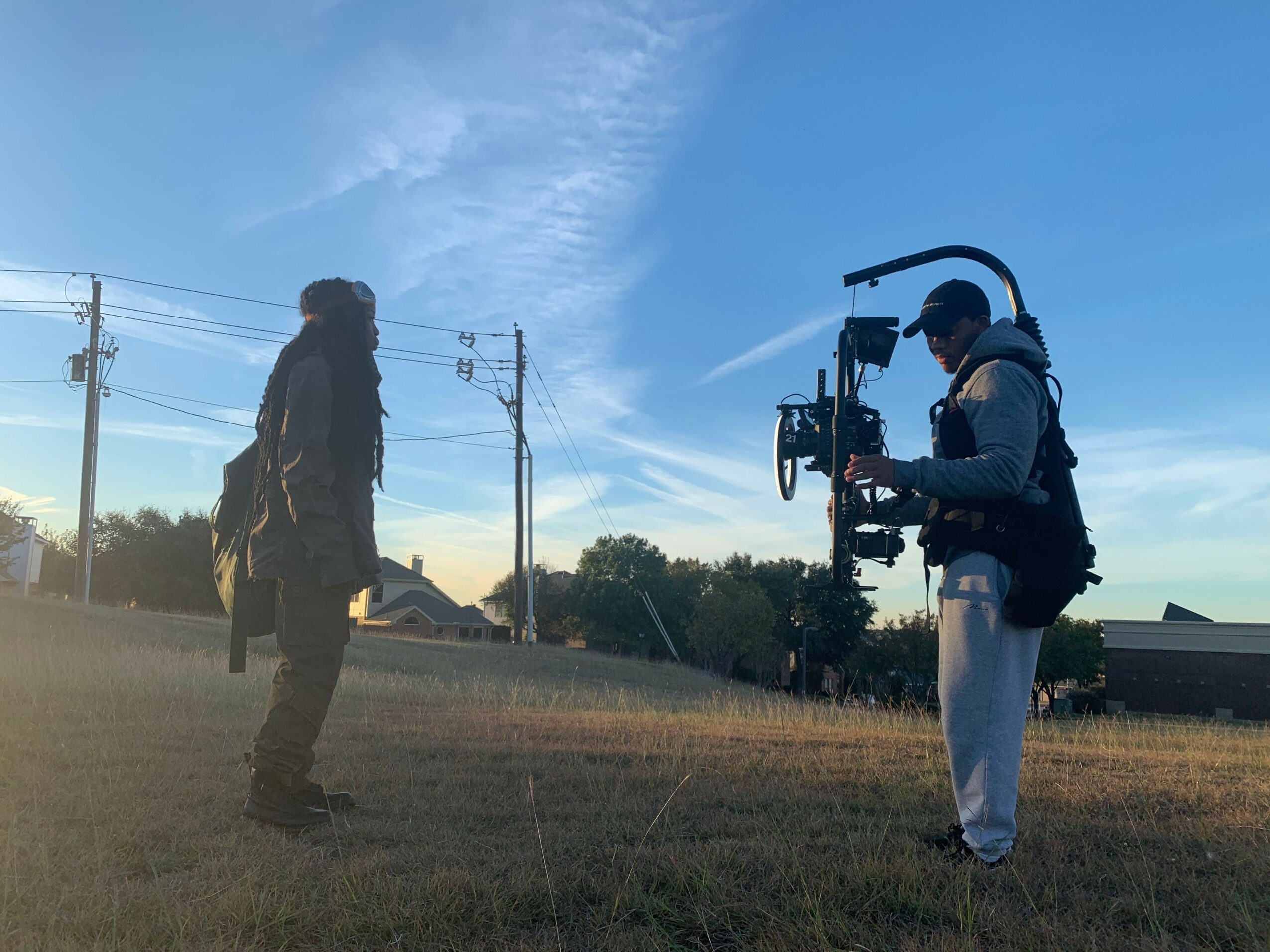 Person operating camera equipment outdoors with a person standing nearby, under a clear sky.