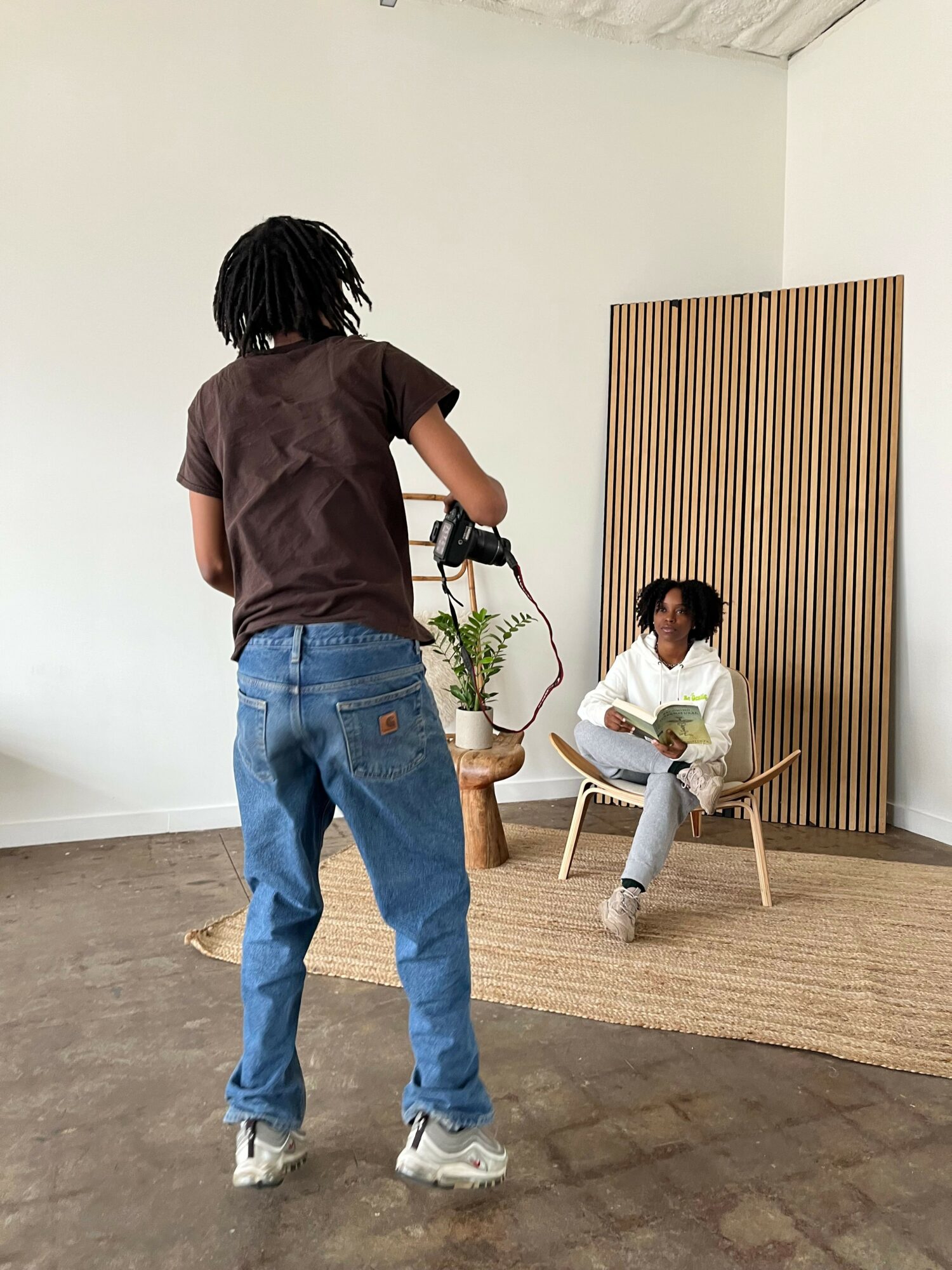 Person with dreadlocks taking a photo of a woman sitting on a chair with a dog nearby, in a room with wooden wall paneling.