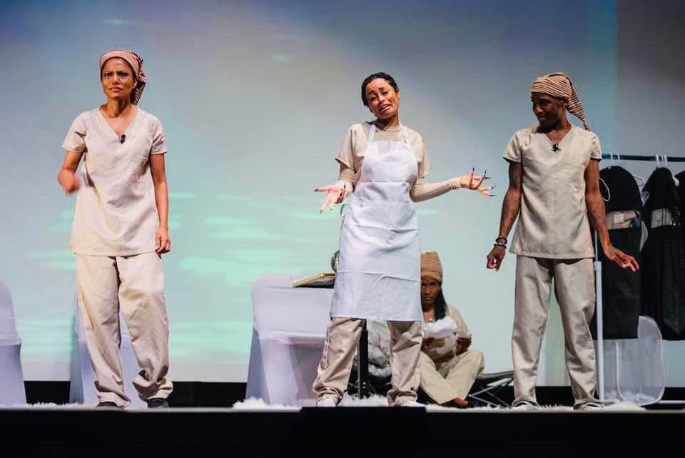 Three women in medical uniforms on stage, one gesturing with hands, two standing, one sitting behind a table, background plain.