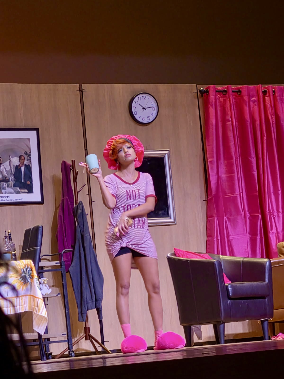 Person with pink curly hair in pajamas and slippers stands on stage, holding a microphone, with a clock and curtains behind.