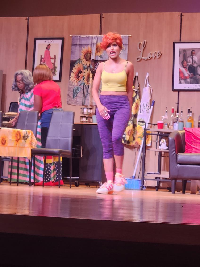 Stage scene with woman in yellow top and purple pants, colorful background, and people in the background.