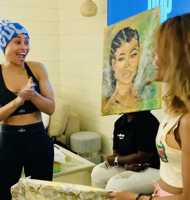 Three women in a room, one smiling and clapping, another with a painting of a woman, and a third partially visible.