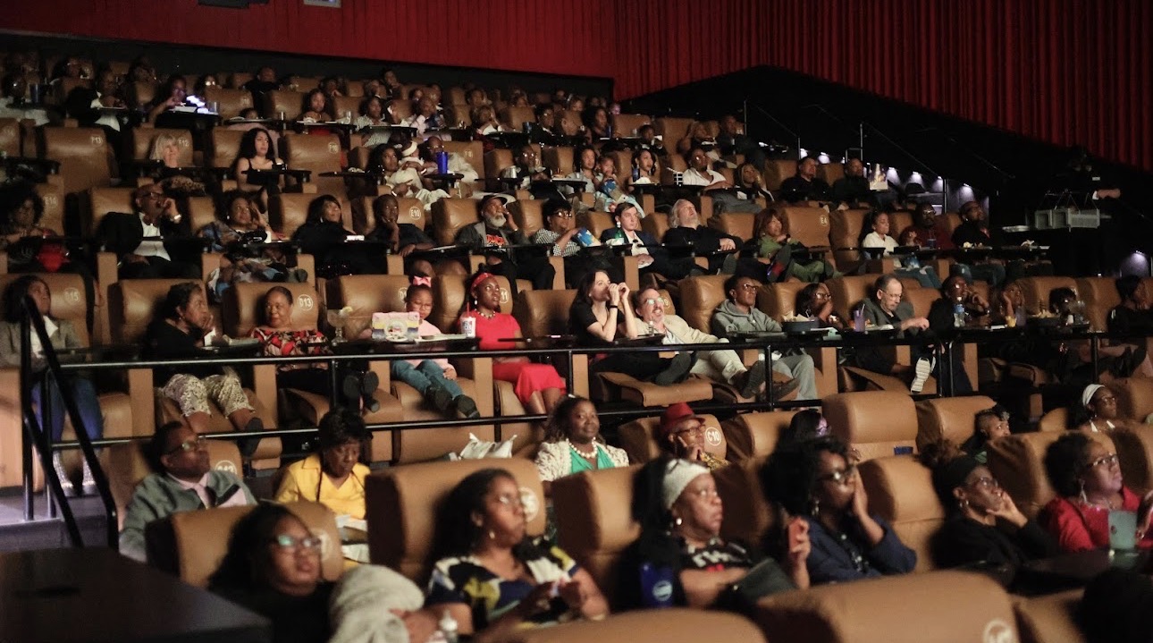 Audience seated in theater, watching a performance or presentation, with many people wearing masks.