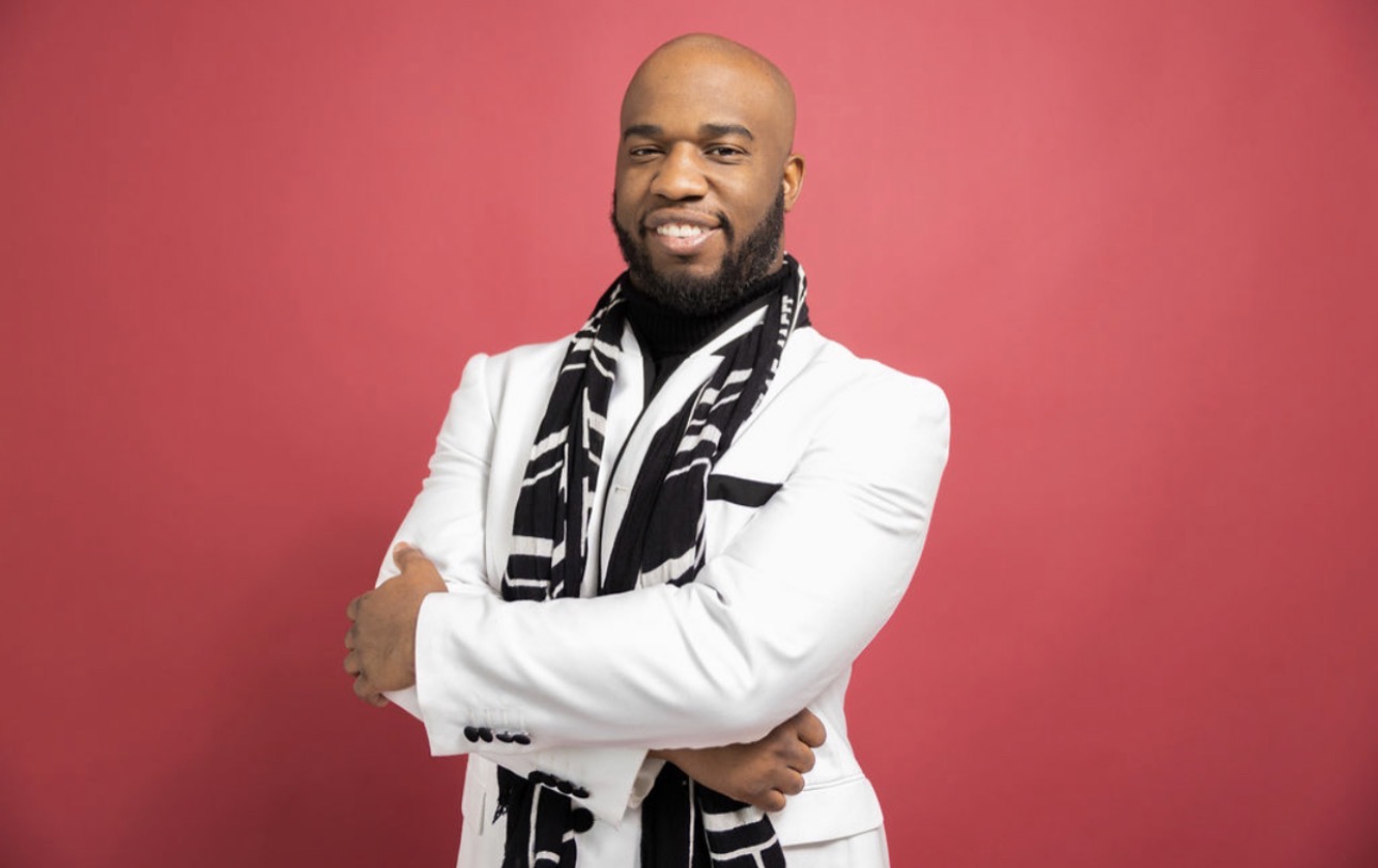 Man with a beard wearing a white blazer and a black and white scarf, smiling against a pink background.
