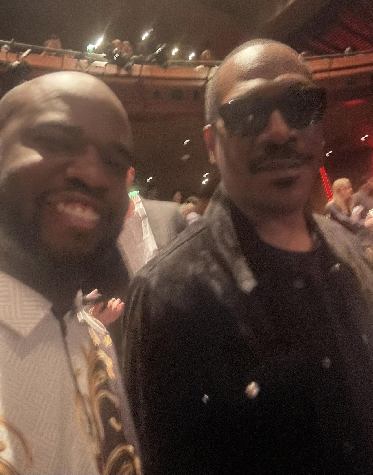 Two men posing together in a dimly lit indoor setting, one smiling and the other wearing sunglasses and a leather jacket.